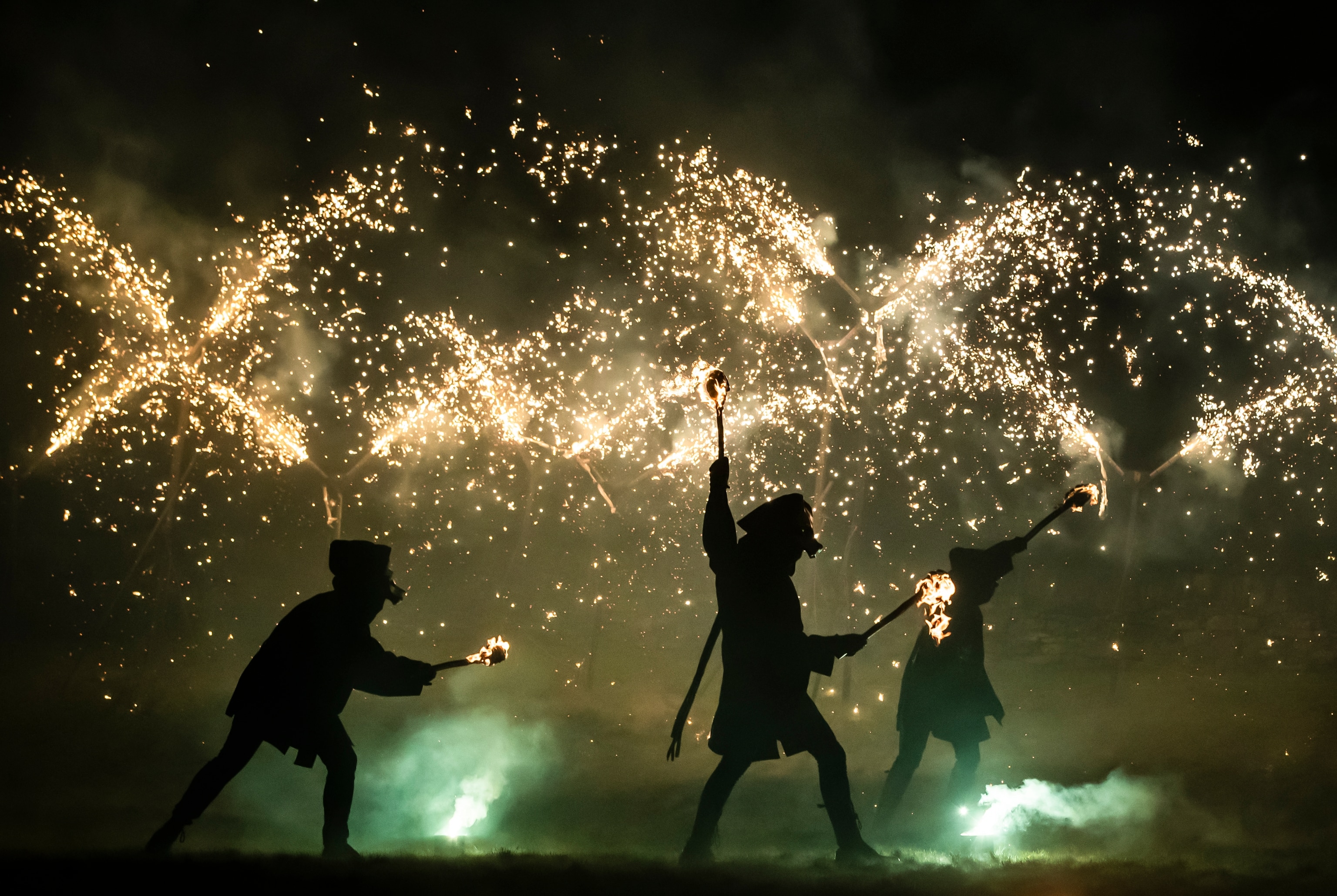 Three dancing figures holding torches are silhouetted against a backdrop of sparks, green flares at their feet