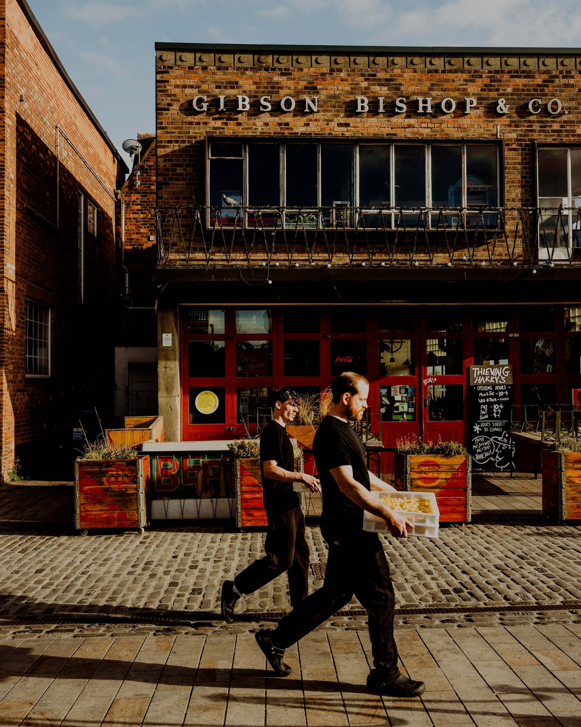 Hull, United Kingdom — July 2025: Scenes from Hull’s maritime heritage and community, showing a view along Humber Street with Thieving Harry’s café and the former Gibson Bishop & Co. building, as two people walk past carrying fresh pasta.