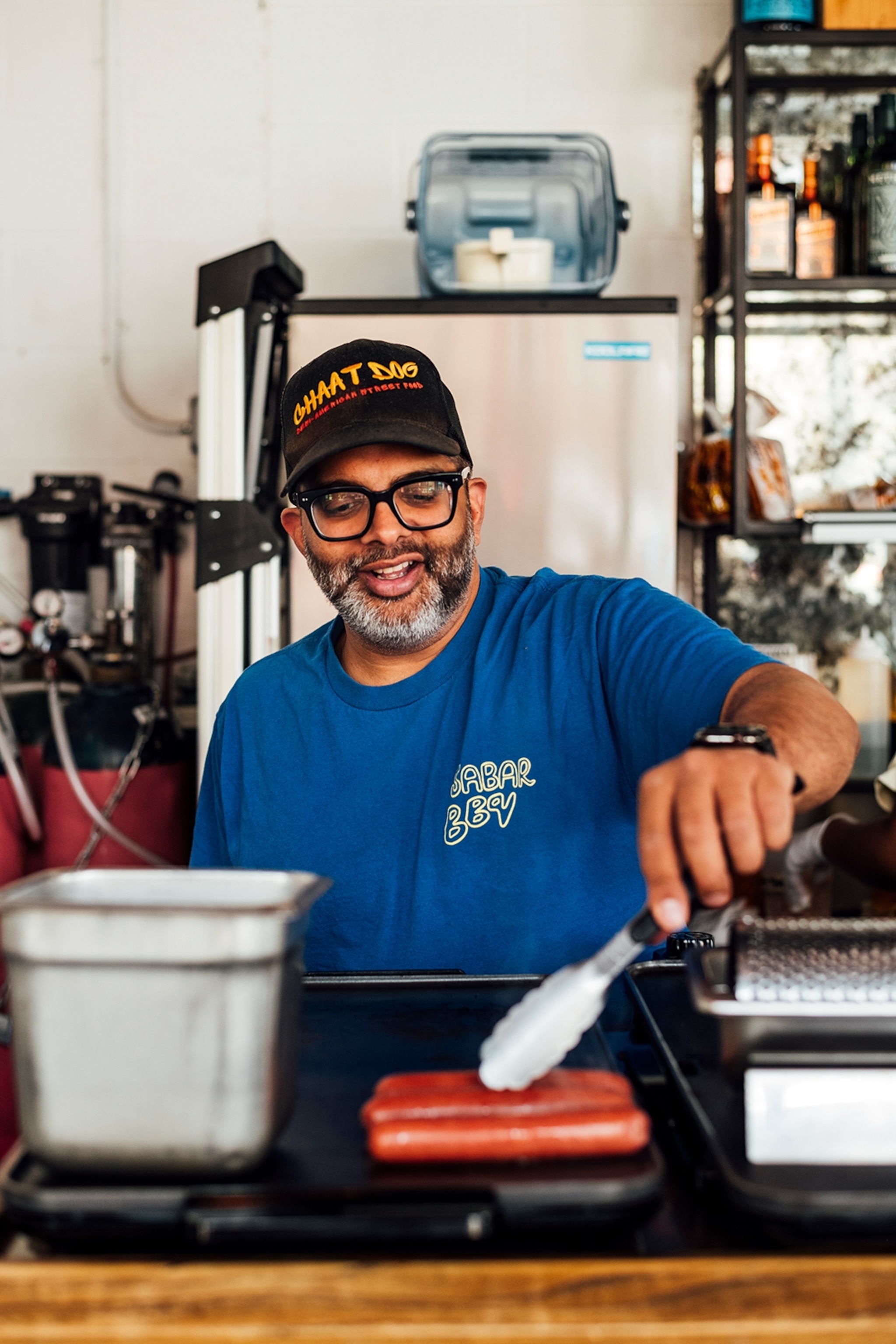 A friendly chef in a simple T-shirt, flipping hot dogs on a grill in front of him.