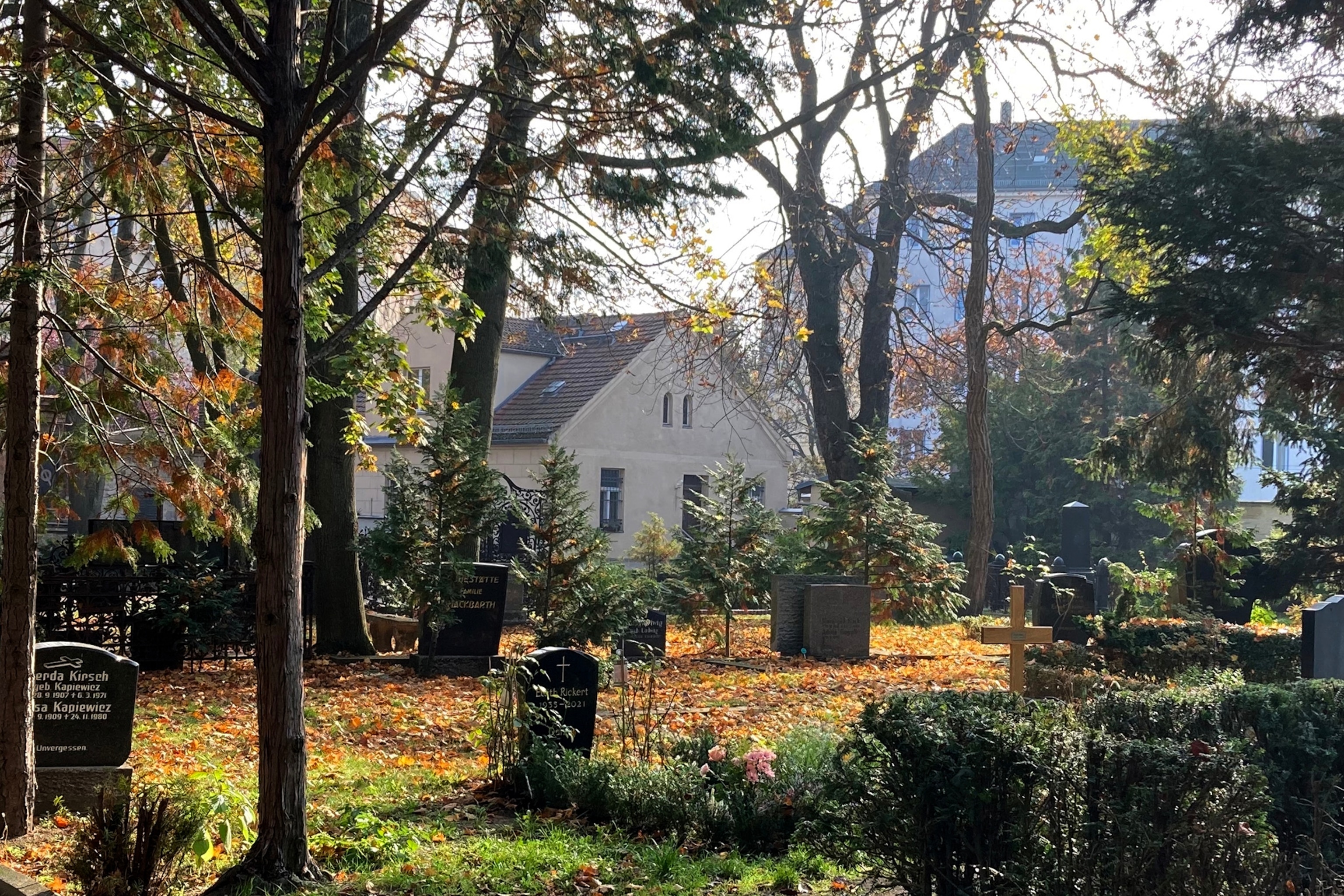 A graveyard in autumn with trees in between the gravestones and a house in the distance.