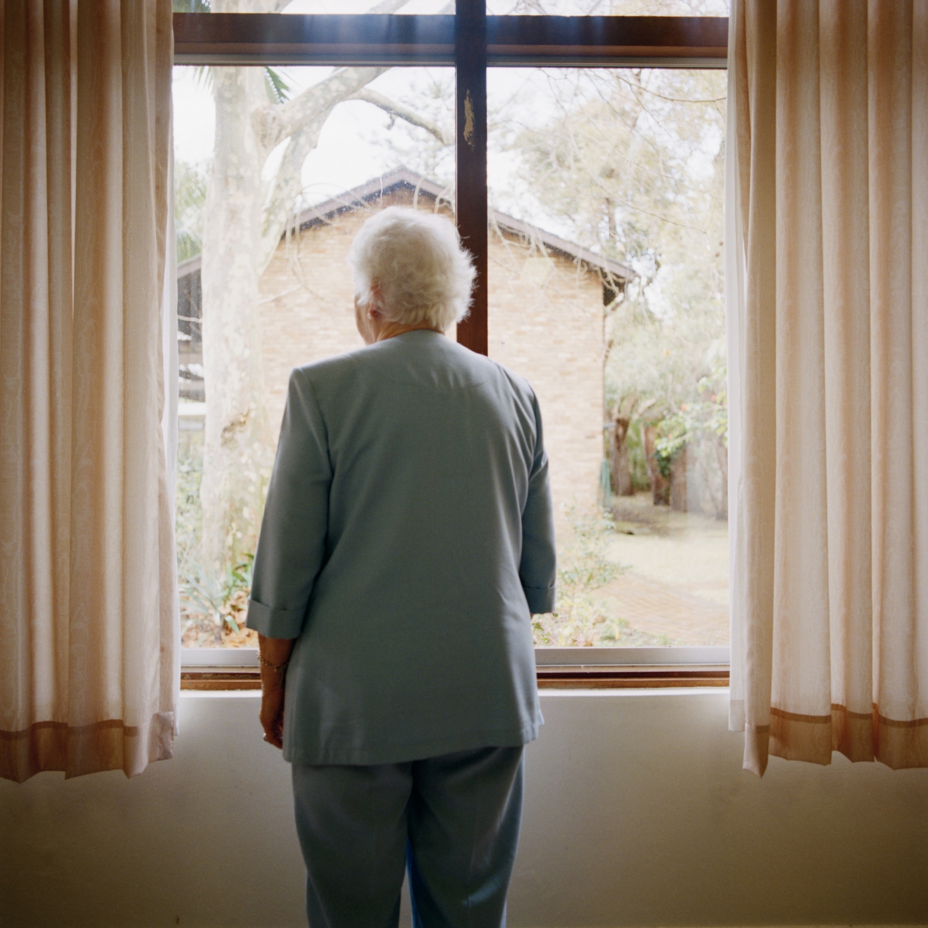 An elderly woman by herself, seen from behind as she looks out a window