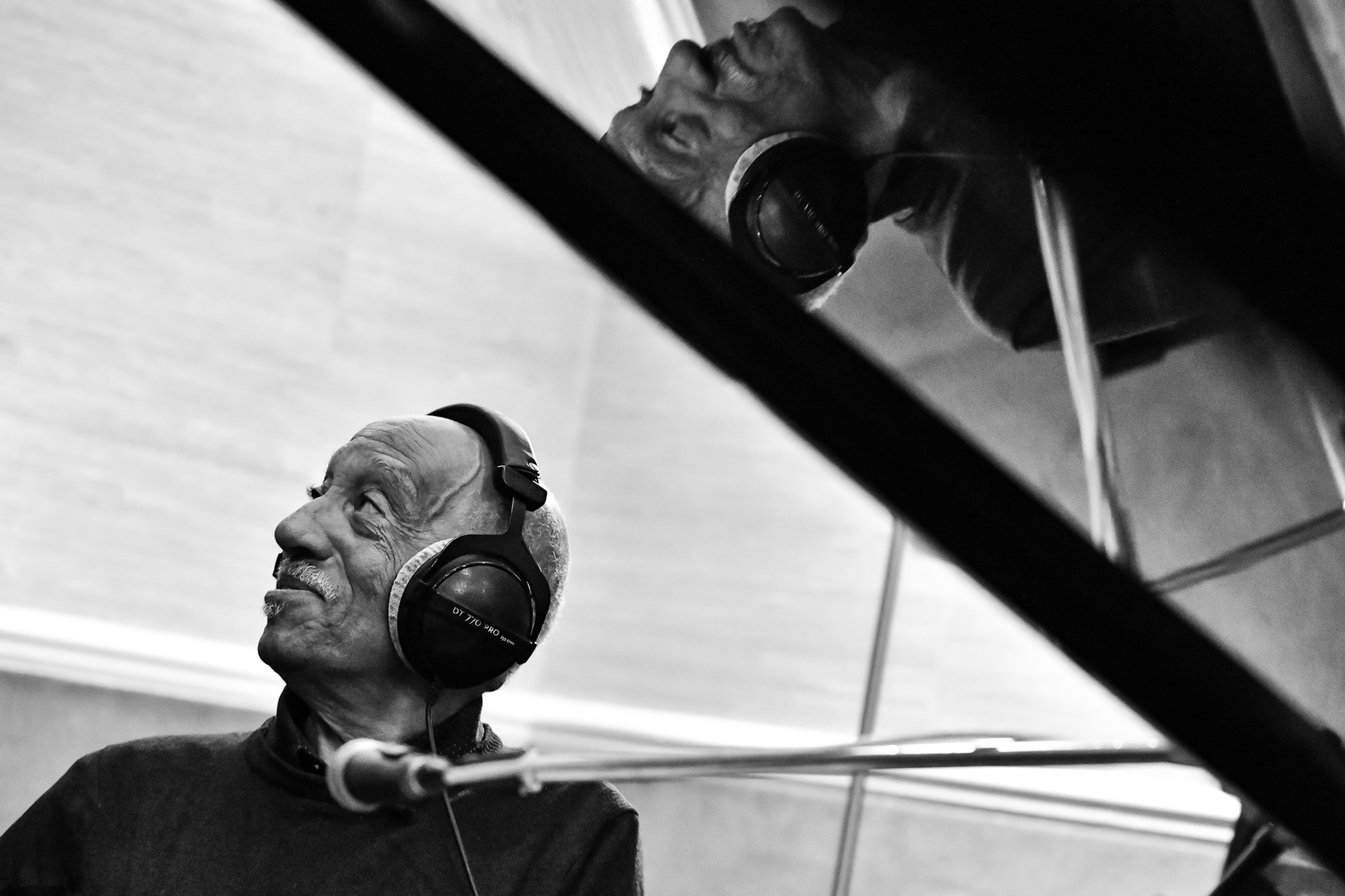 A black and white photograph shows a man at a grand piano with headphones on his reflection is in revers above him.