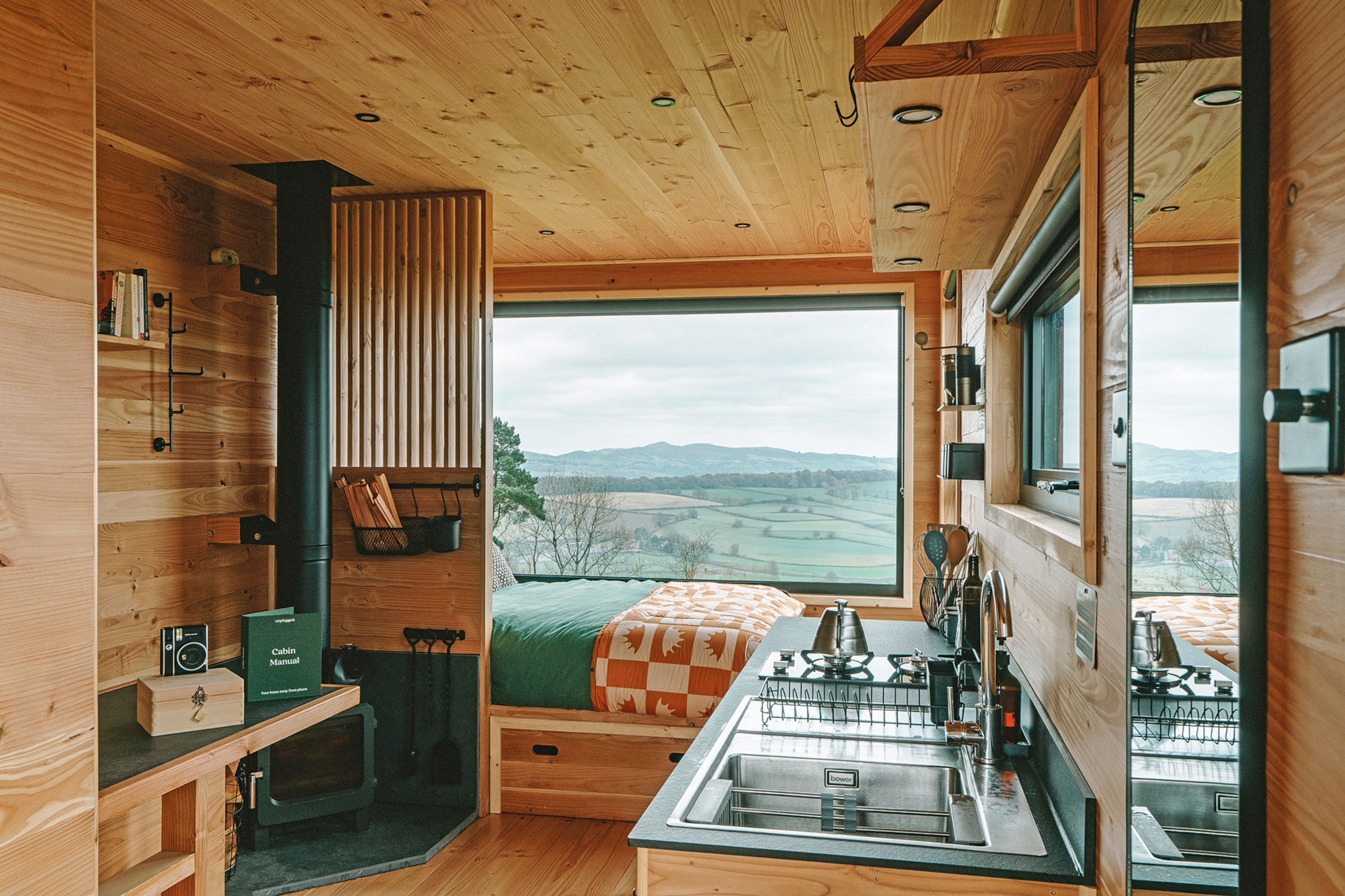 The interiors of a modern, wooden cabin with a small kitchen, chimney and bed in front of a wall-wide window with views of a hilly landscape.