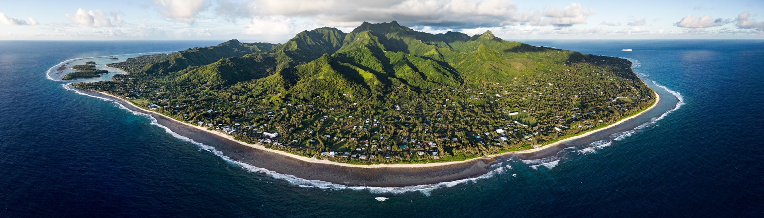 Aerial view of the island with green mountain in the middle, human settlements along beaches, surrounded by blue sea and white water of the surf zone.