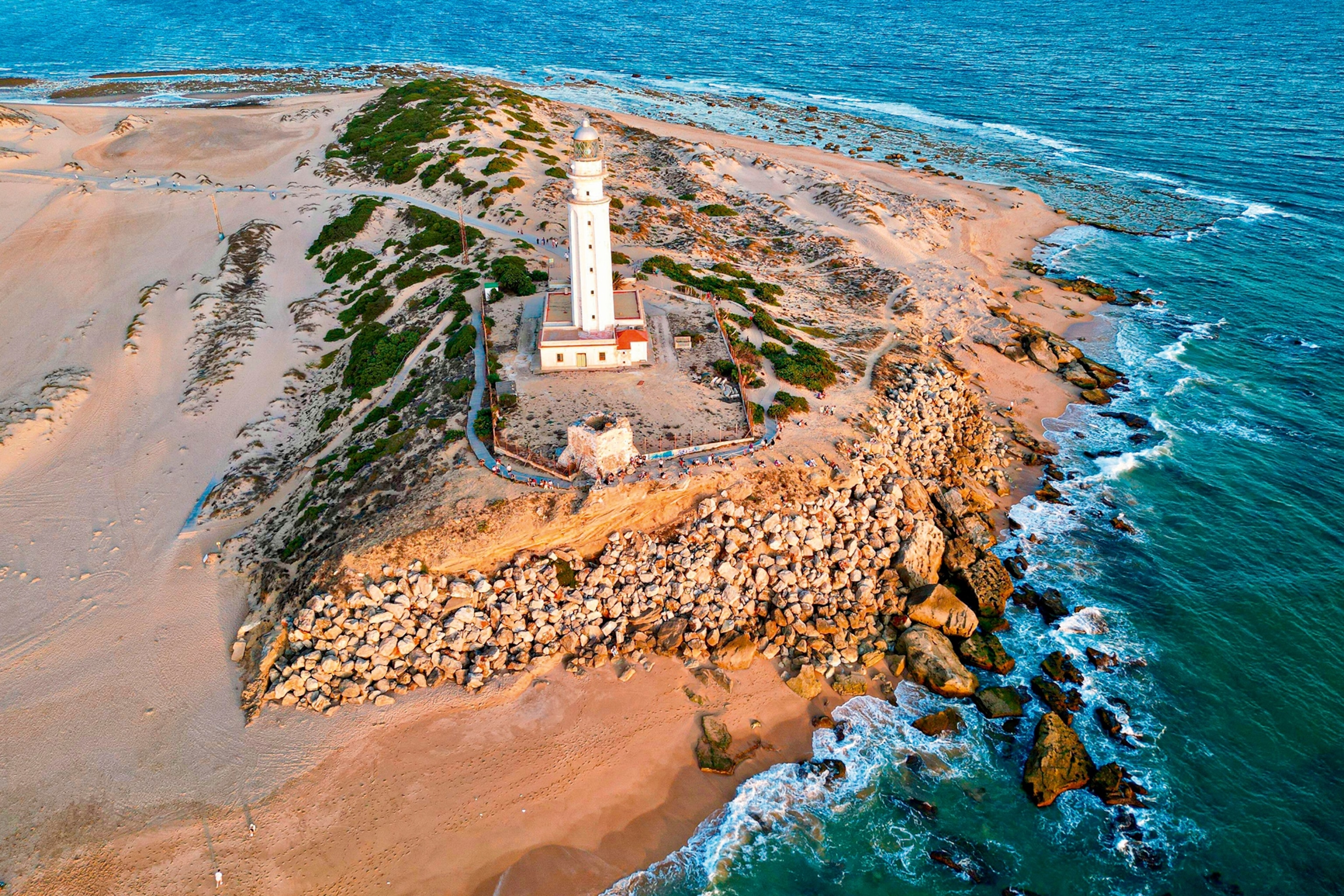An aerial shot of a windy, rock-lined coastline with a lighthouse at the edge.
