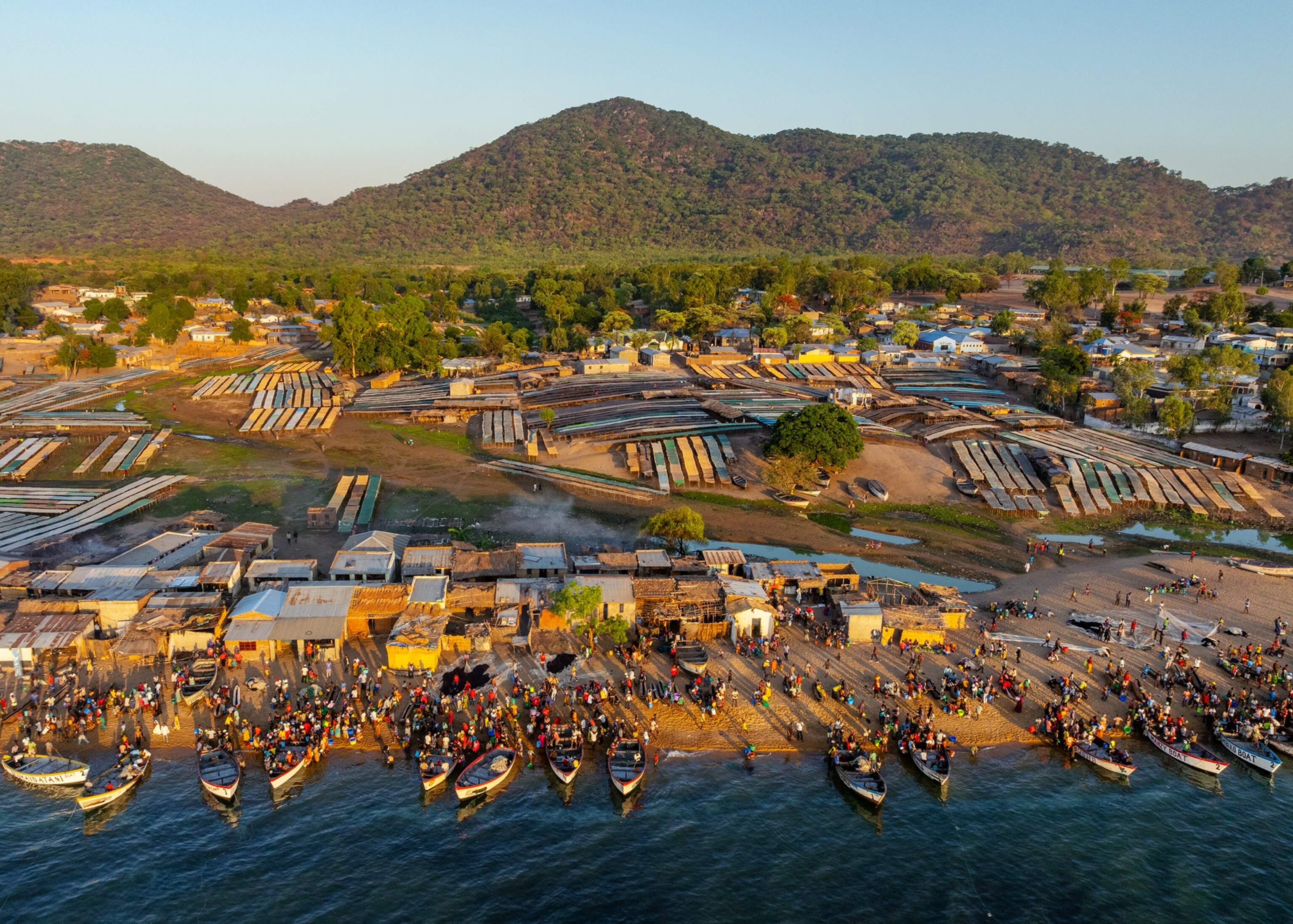 An aerial view of a busy lakeside community shows rows of boats lining the shoreline, crowds gathered along the waterfront, and a network of buildings and market structures stretching inland toward green hills and mountains.
