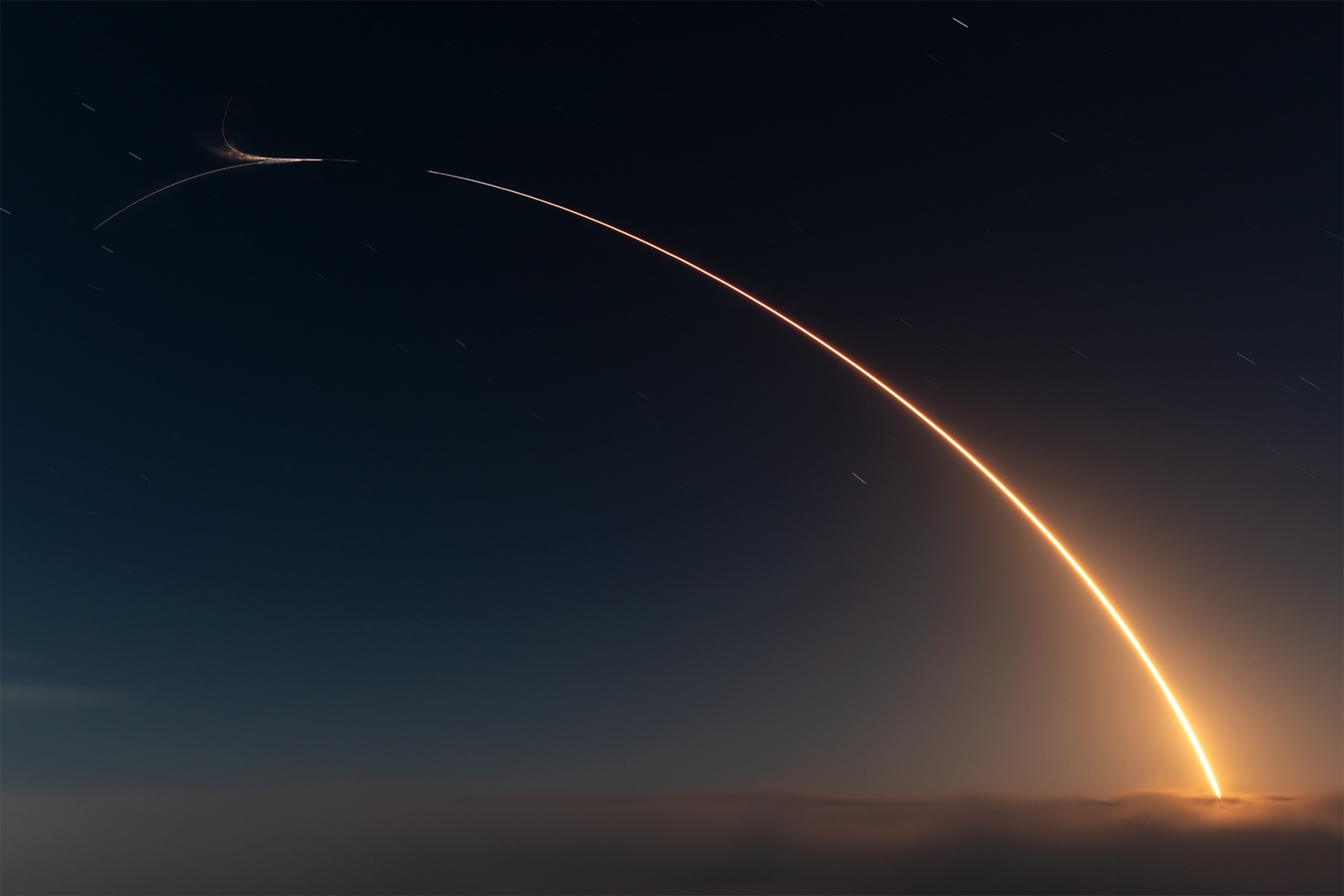 A glowing rocket trail arcing across a dark, starry sky, emanating from a cloud layer