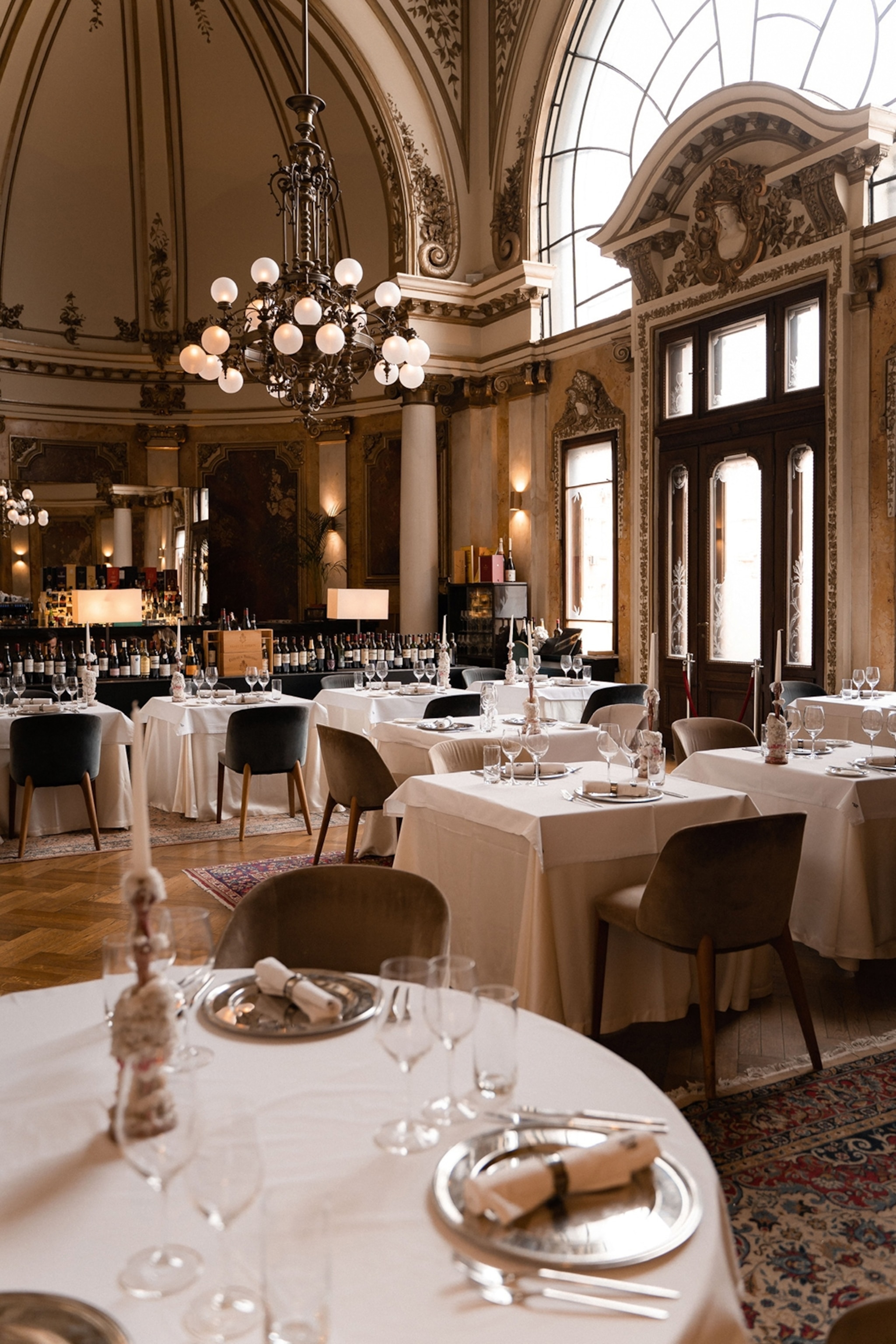 An old palatial restaurant with linen-decked tables, tall windows and decorative features.