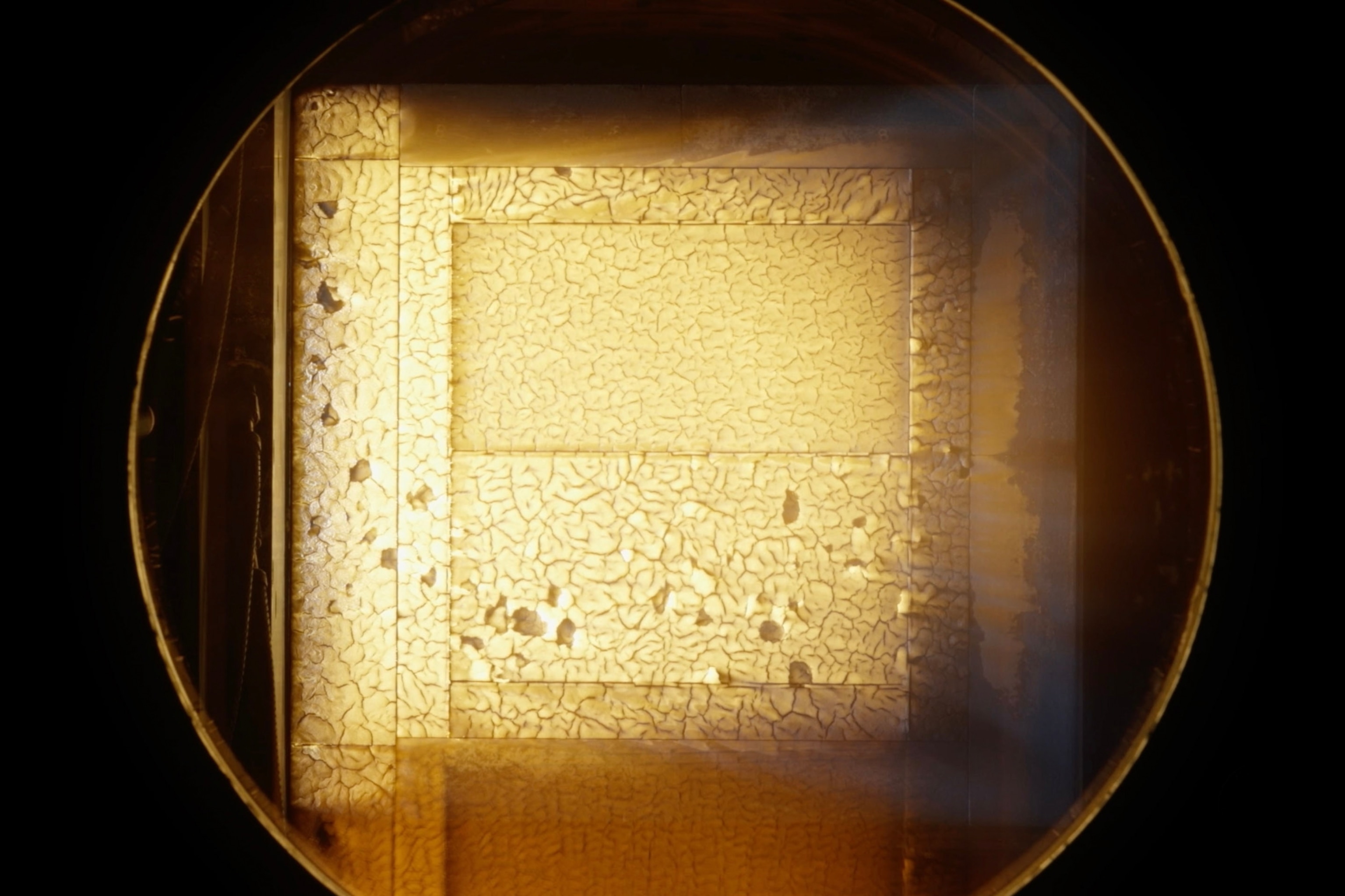 Close-up of a metallic surface under a microscope, showing a textured, cracked pattern with a golden hue