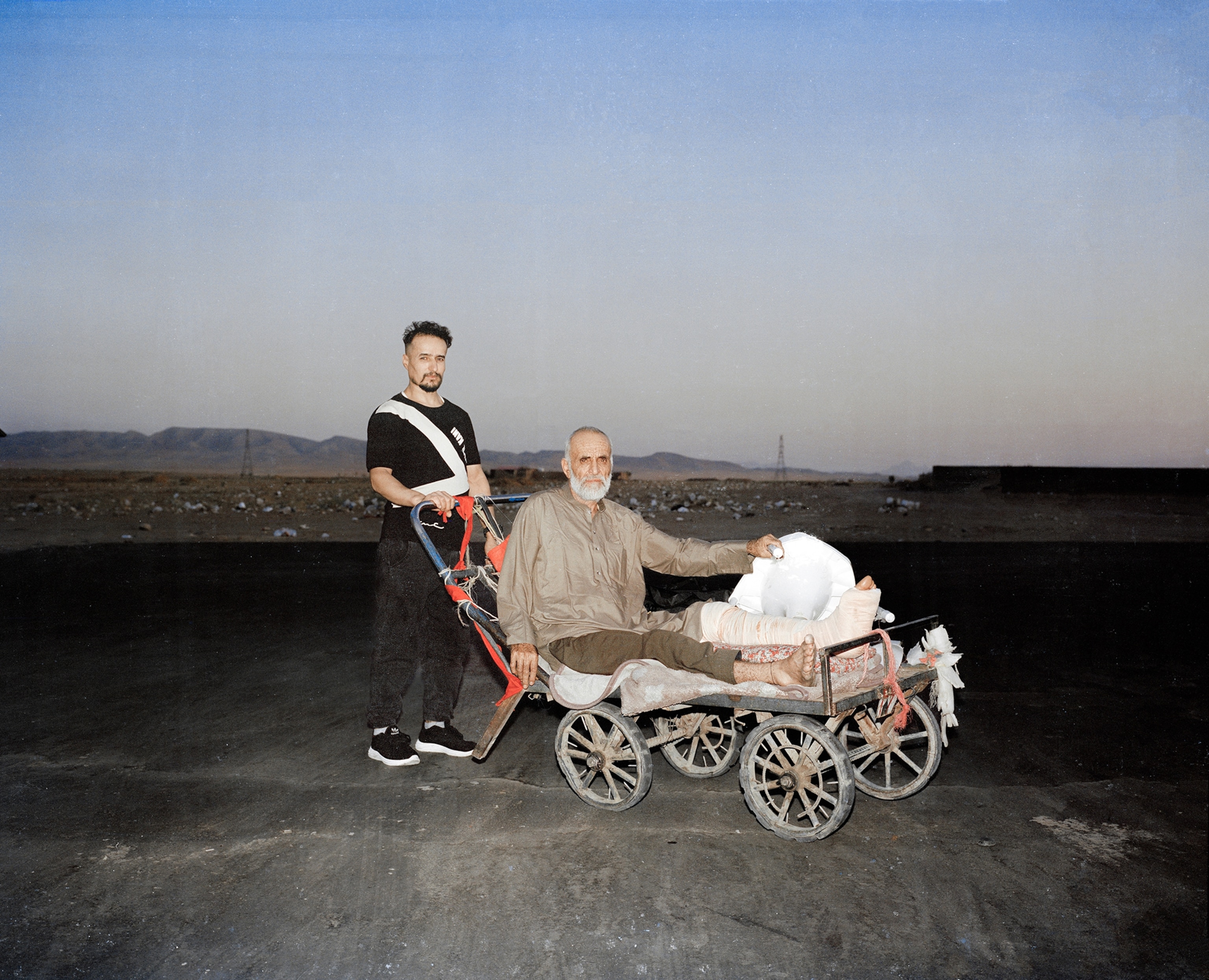 An older man in a makeshit wheelchair with his left leg in a full cast is helped along by a younger man. 