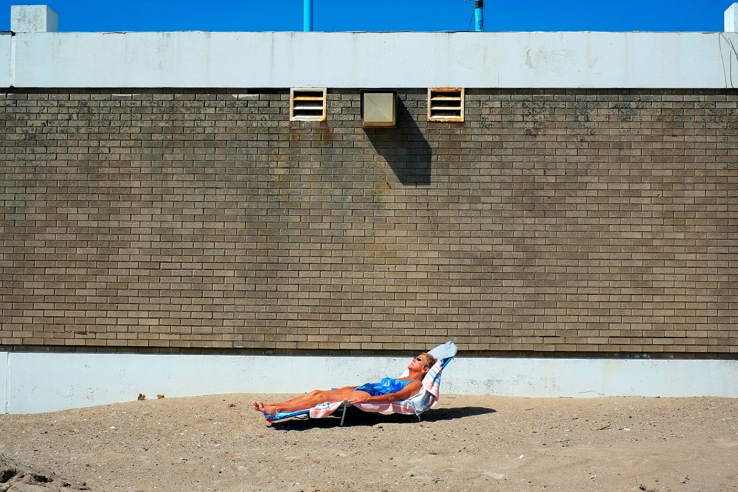A woman sleeps on a chair on a sandy beach in front of a brick wall.