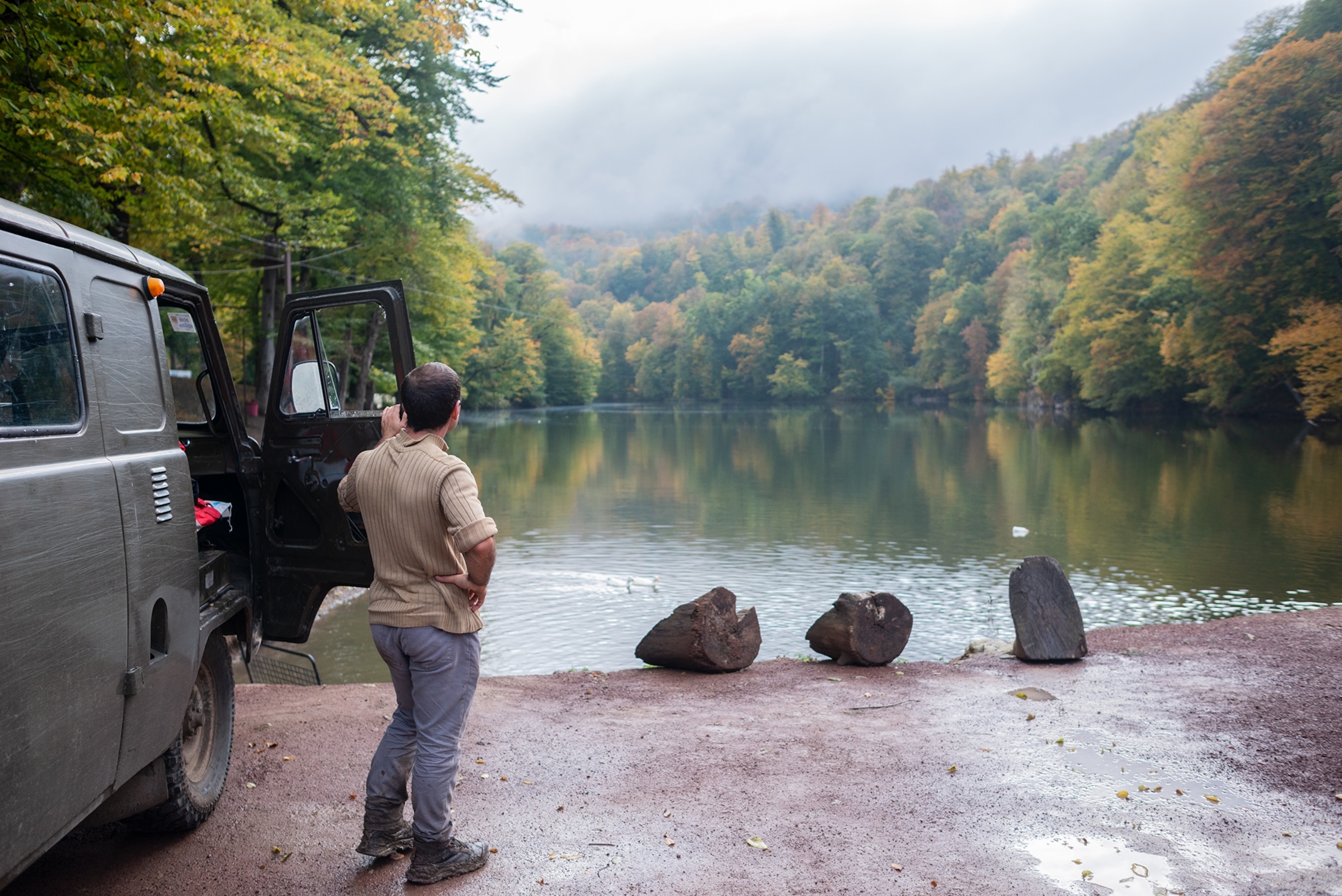 A person stands beside an off-road vehicle at the edge of a calm forest lake, looking across the water toward tree-covered hills and misty mountains in the distance.