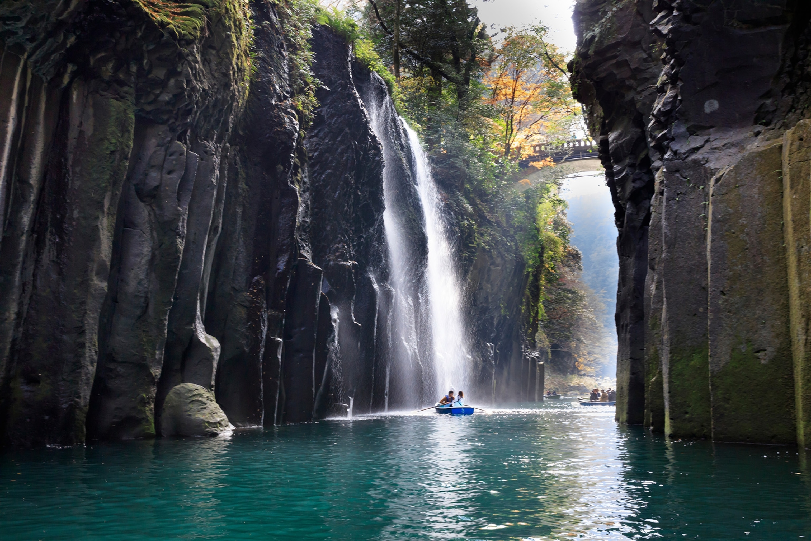 People sit in a rowboat on a river with a waterfall and a bridge in the background
