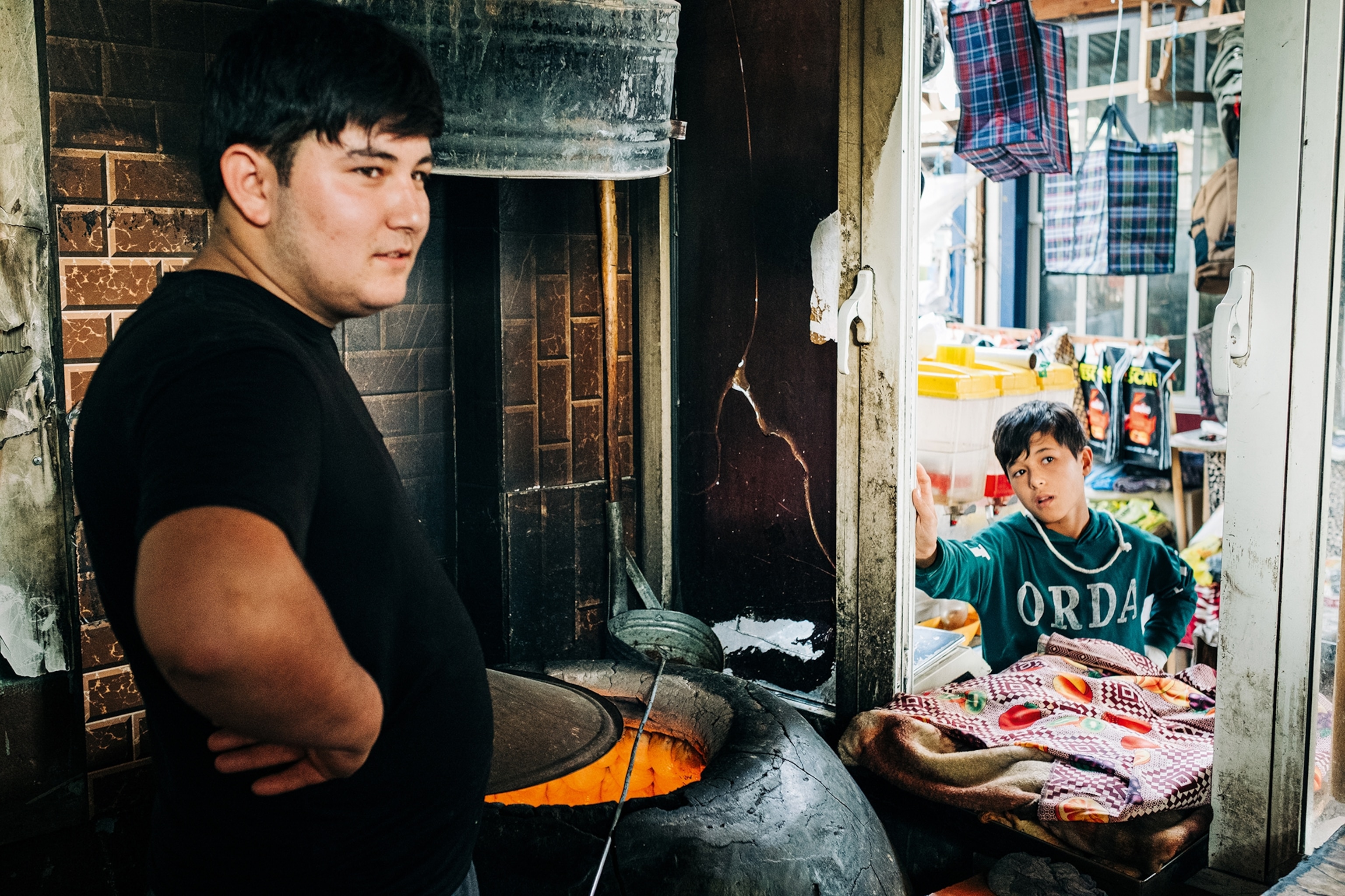 A wide perspective from the inside of a street food stall with a young man waiting for bread to finish in a tandoor oven as a boy customer waits in the stall window.