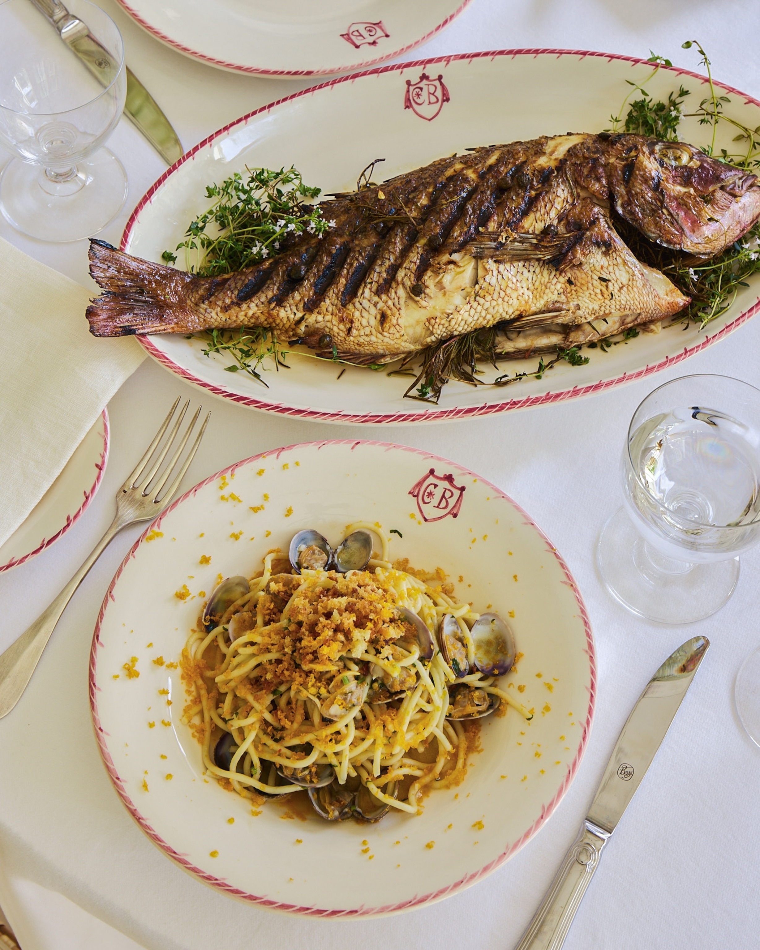 A table set with a grilled whole fish and a plate of clam pasta, surrounded by glassware and silverware.