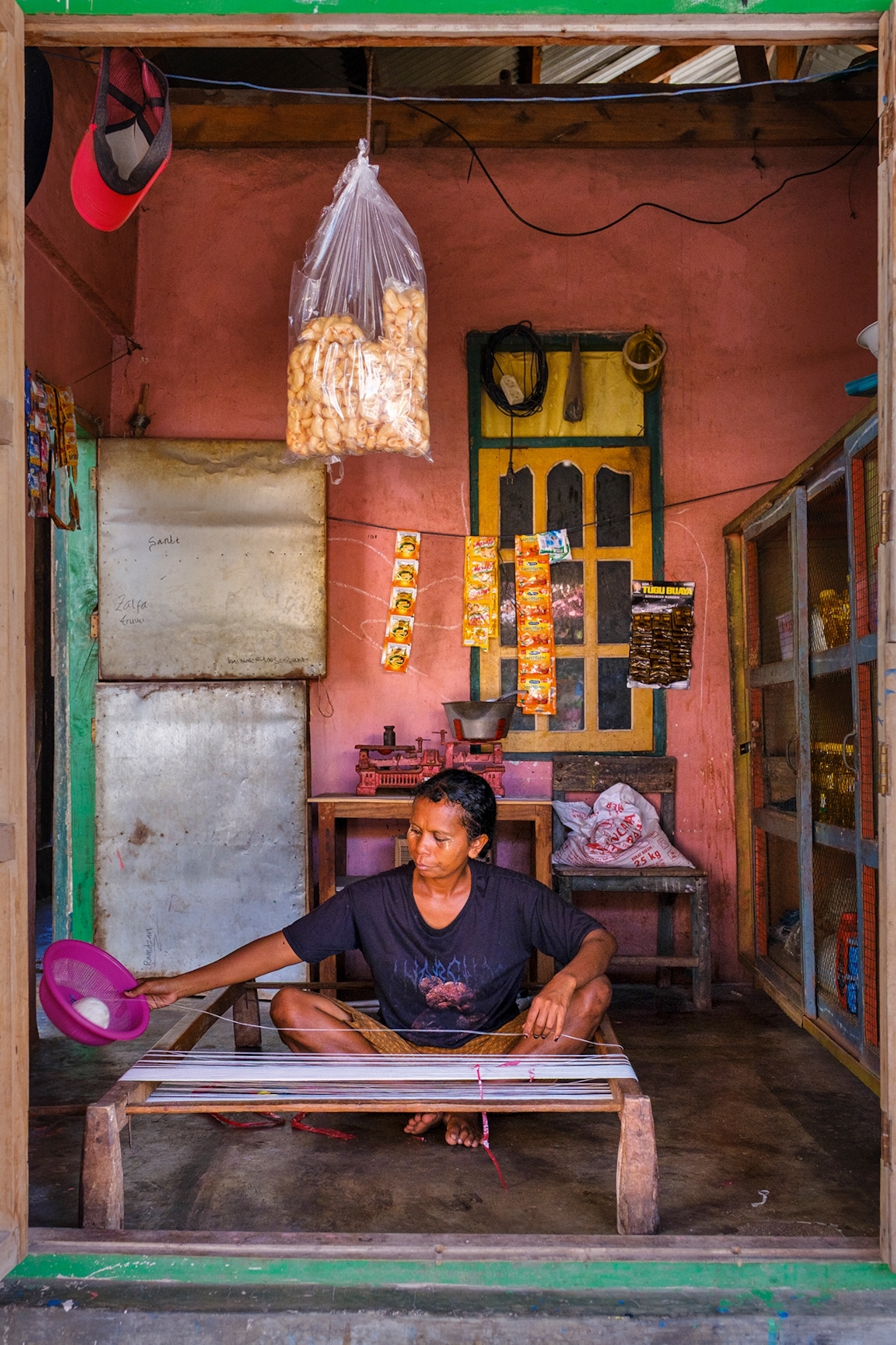 A local Indonesian woman sitting cross-legged on the floor of a simple and weathered room, spinning yarn onto a wooden frame.