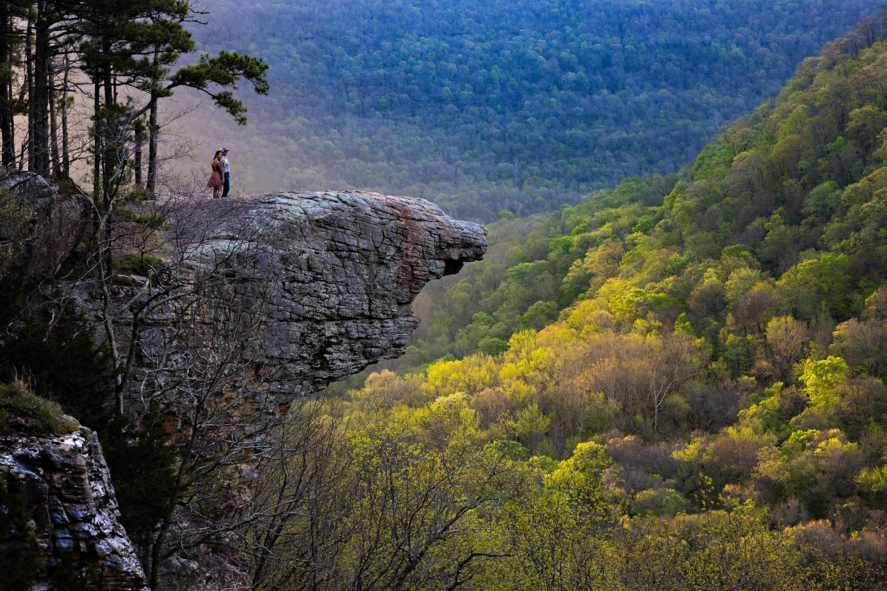 A person stands at the edge of a large rock overlook above a forested valley, with rolling green hills and dense trees stretching into the distance.