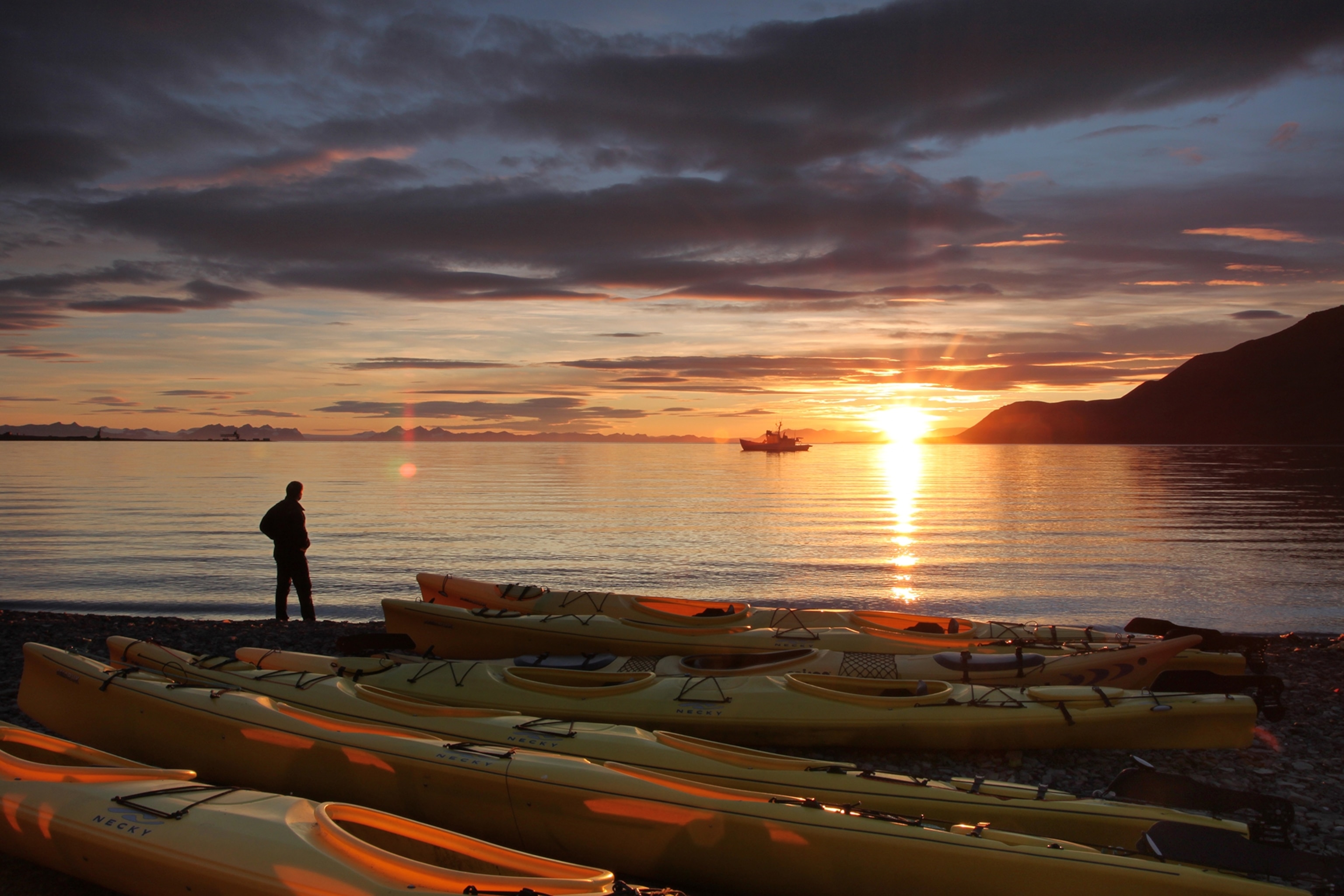 A man stands looking out at the water in Svalbard as the sun sits just above the horizon.