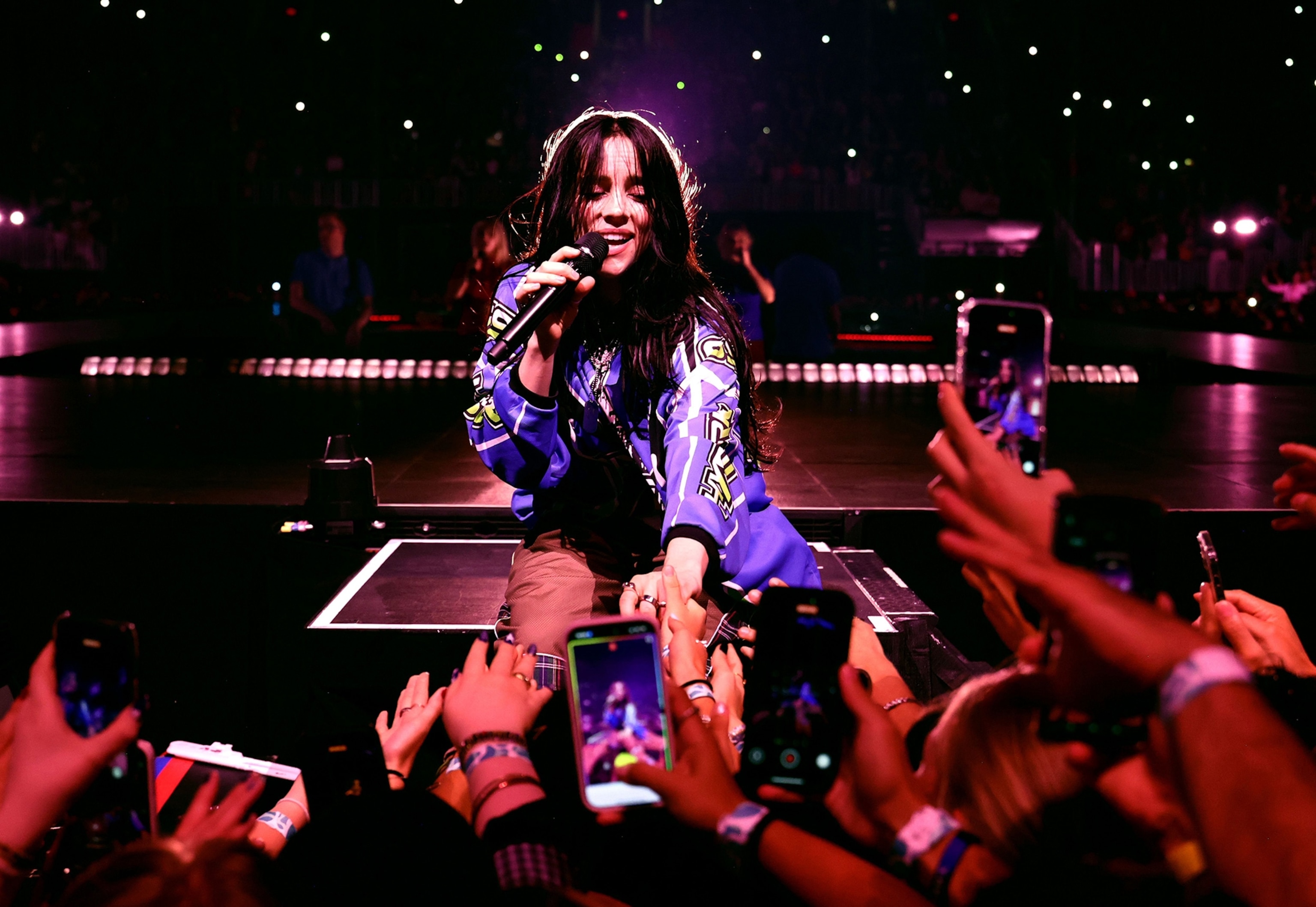 Singer with long dark hair wearing a purple jacket on stage is holding a microphone and engaing with the audience.