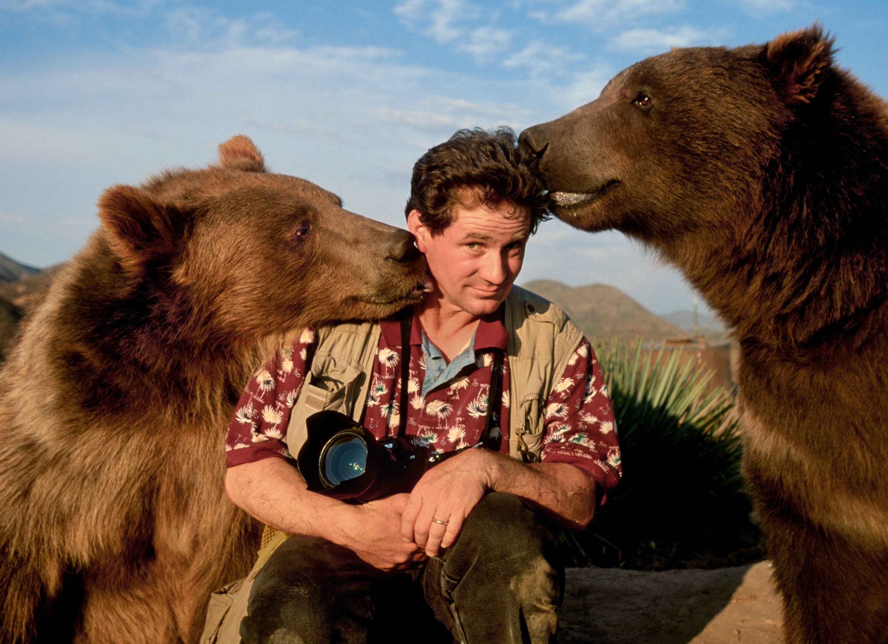 Joel sits with a camera in his hand as two grizzly bears sniff his head.