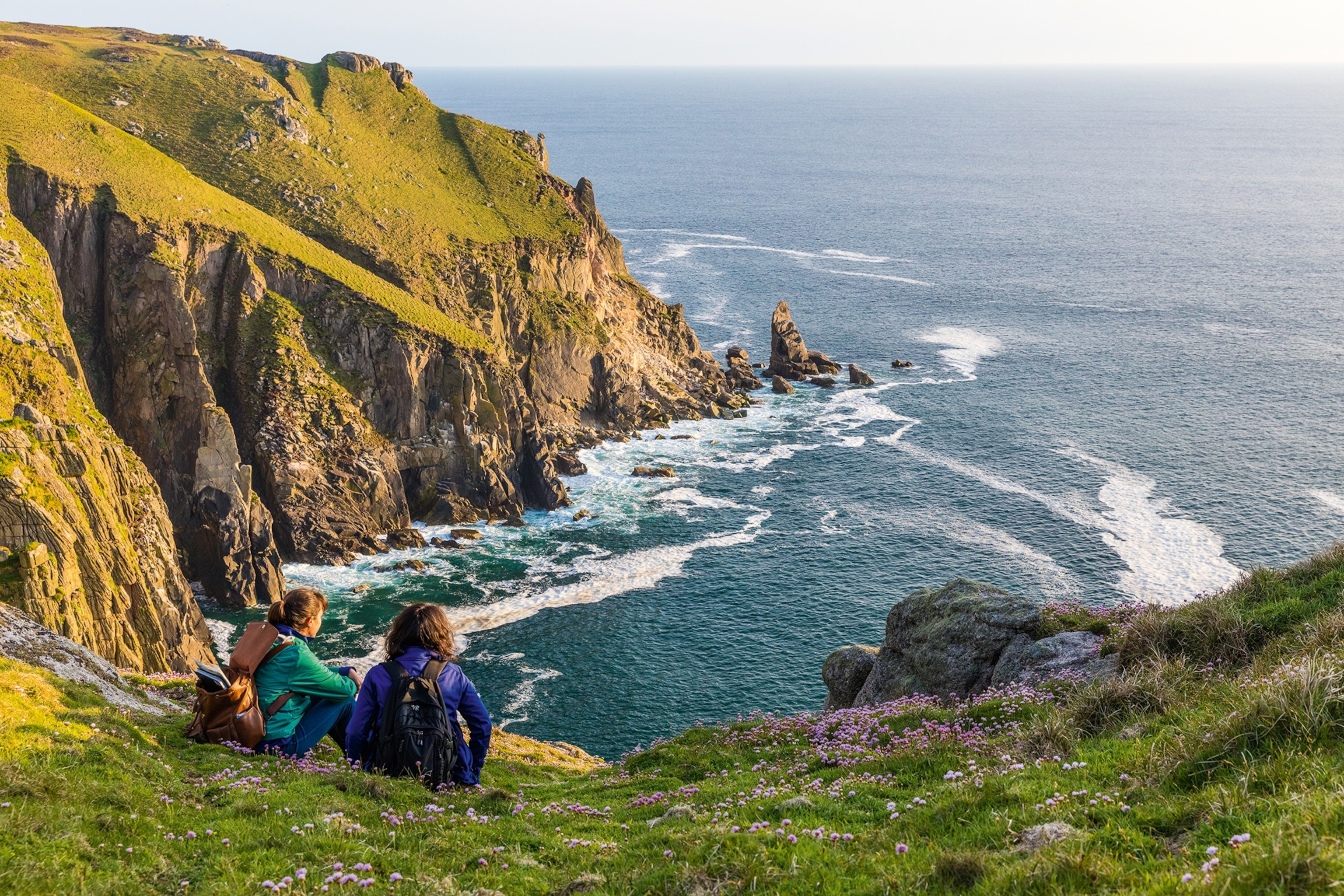 Two women sitting on the edge of a cliff overlooking the rugged coastline ahead.