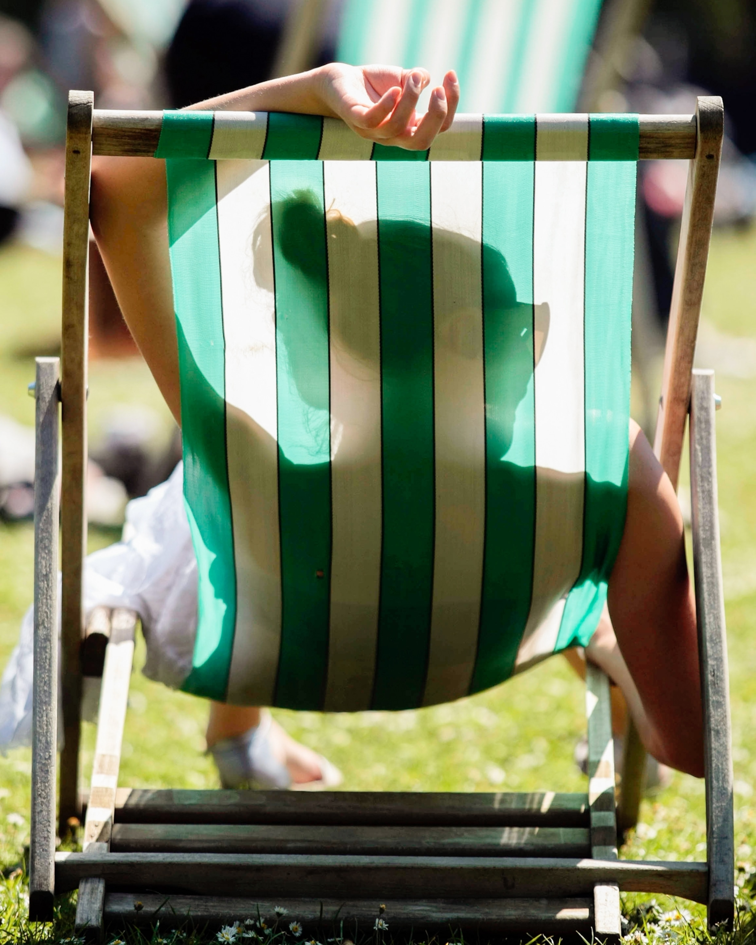 A woman seen from behind lounging in a striped green deck chair on a sunny day in a park, arm draped over the top