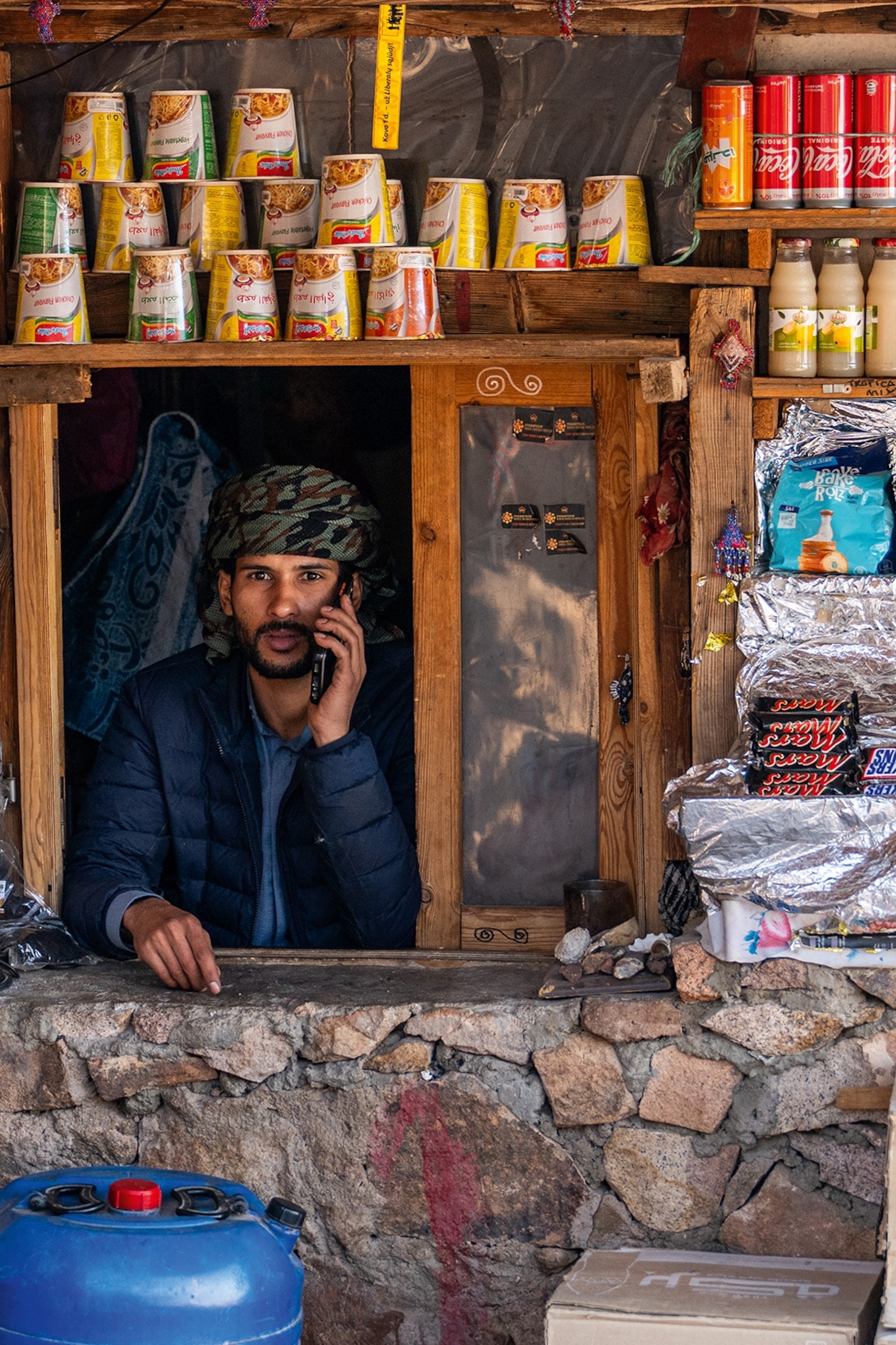 A man sits at the open window of his shop, surrounded by stocked shelves.