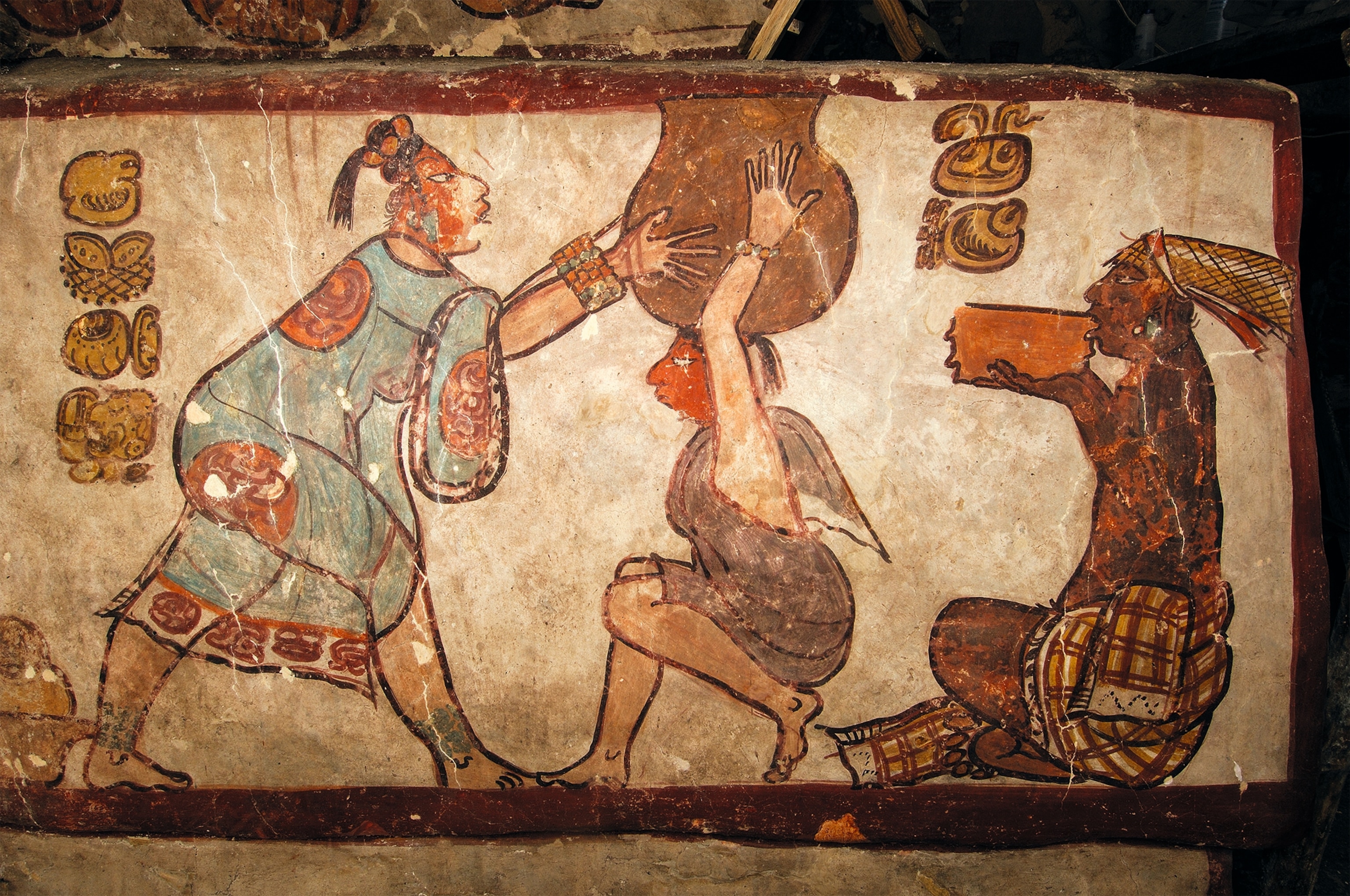 A woman in a translucent blue dress (huipil) decorated with hieroglyphs reaches out to and holds a large vessel on the head of another in less elaborate clothing.