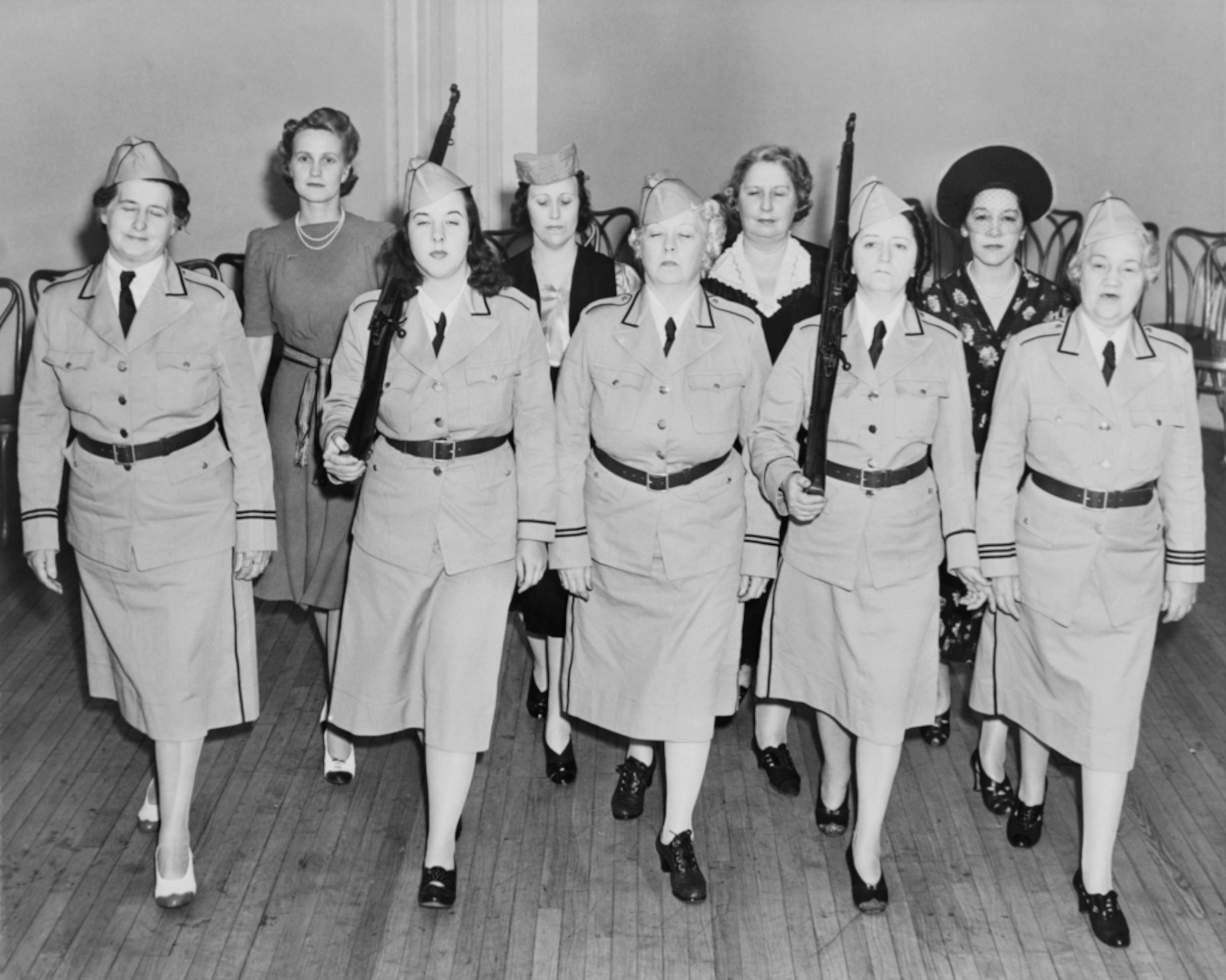 A black and white historical photo of a group of women in uniform, standing in formation indoors, holding guns