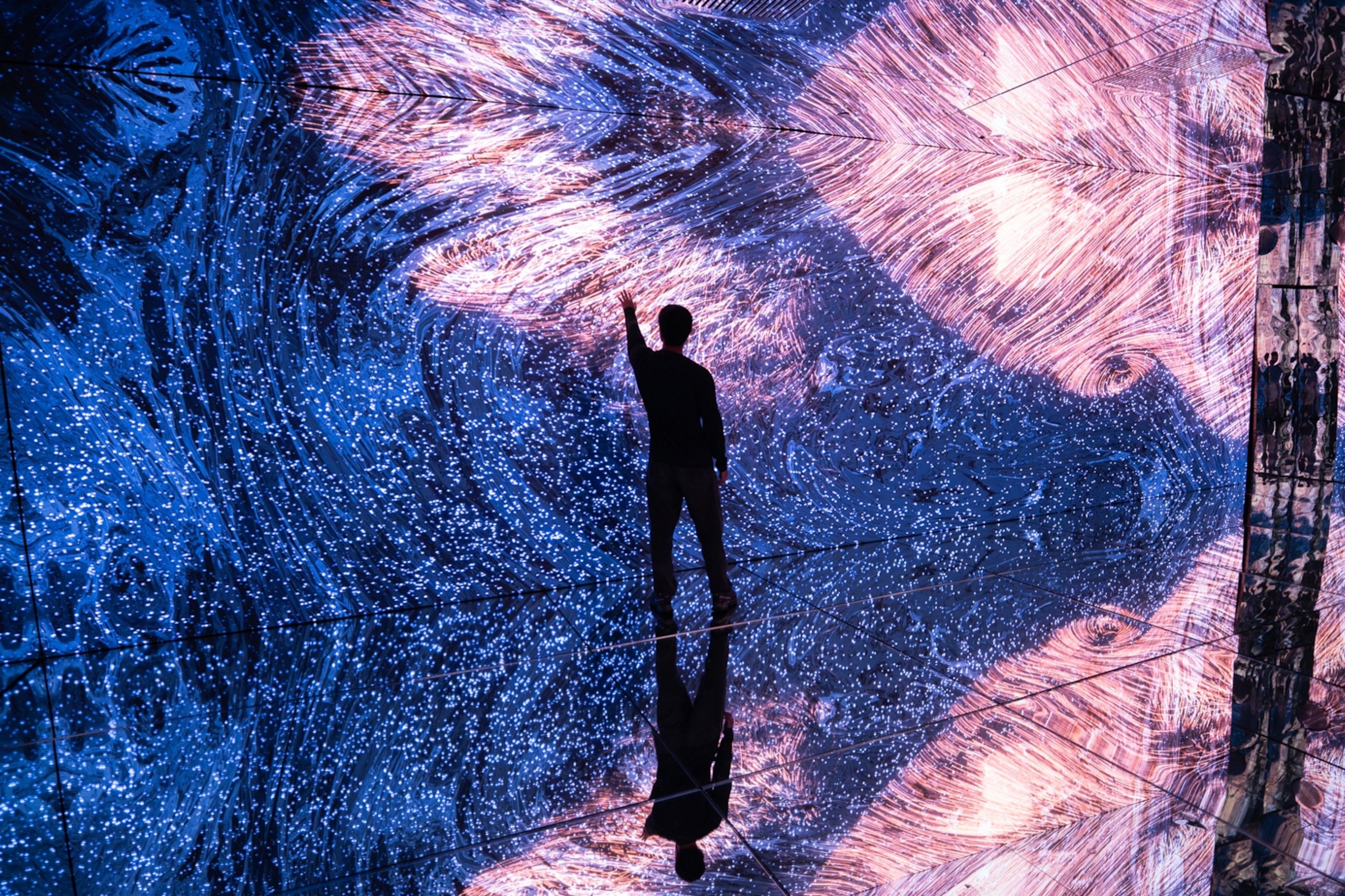 A person standing in an immersive exhibition space with lights bouncing around all walls in the dark.