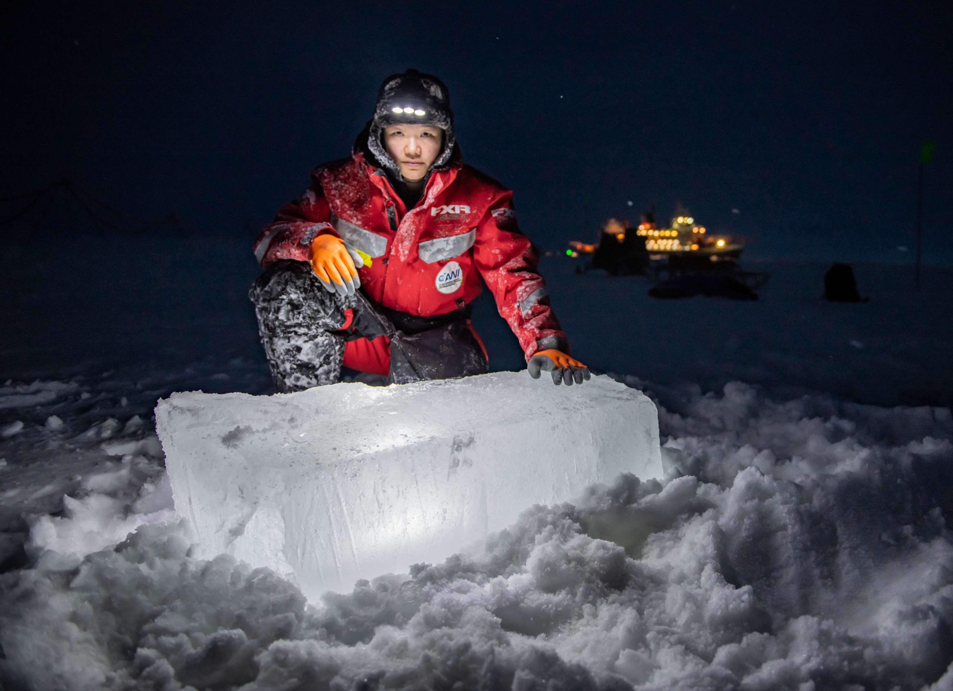 In polar darkness, woman an red thermal jacket and hat with lights is looking in the camera while standing on one knee behind a large rectangular block of ice which reflecting lights to her face.