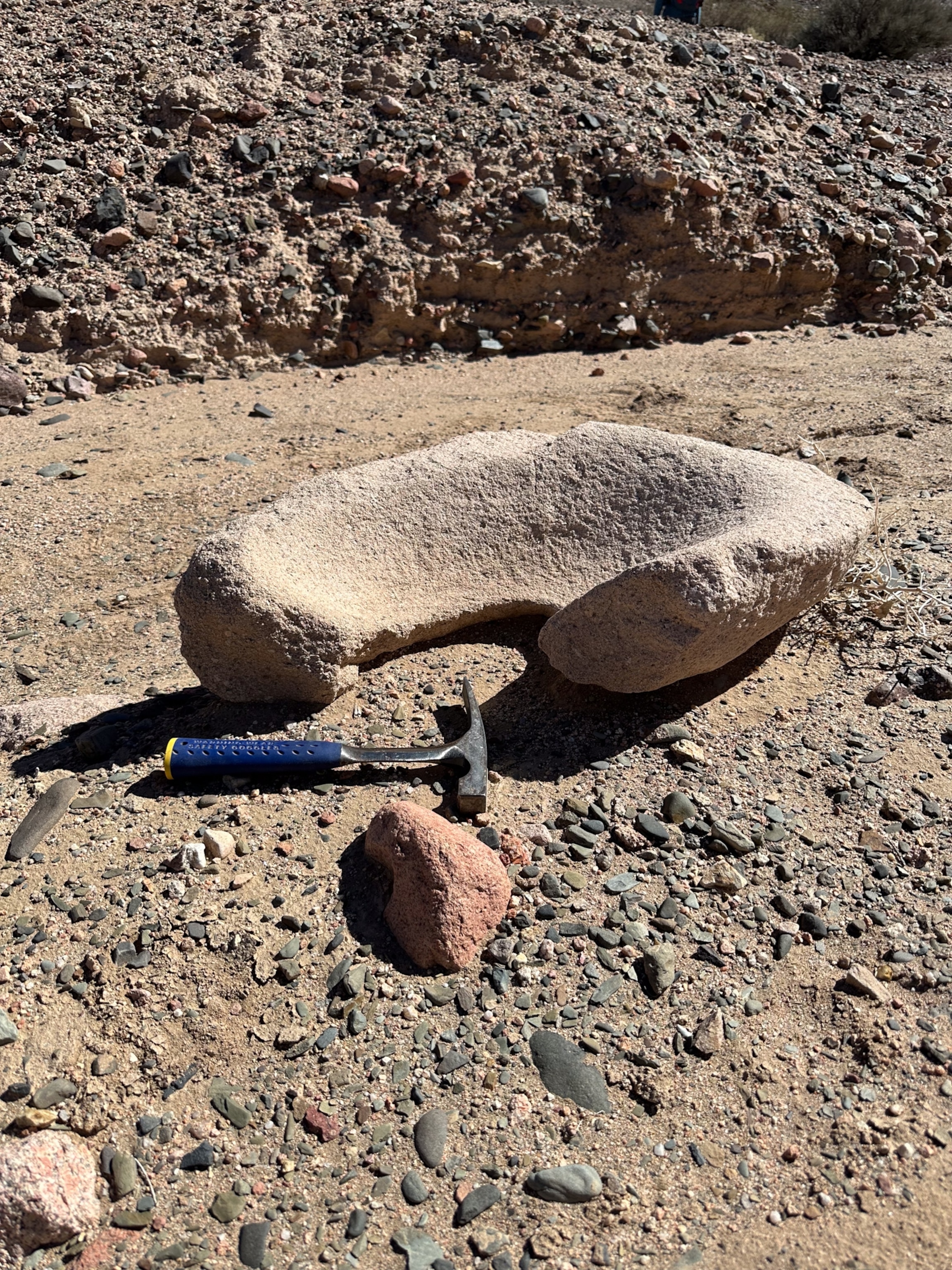 A U-shaped stone lies on rocky ground next to a small red rock and a geological hammer