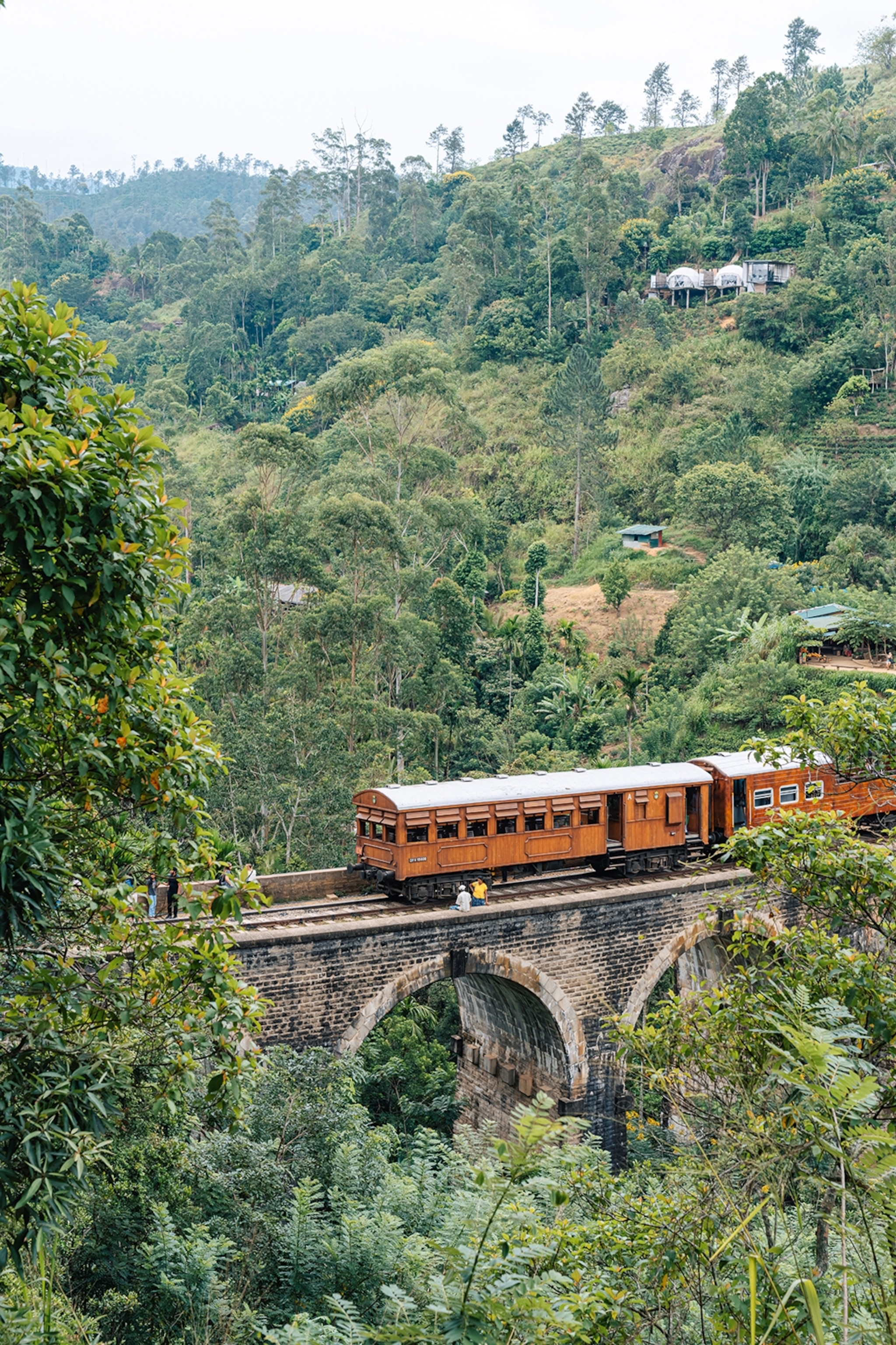 A wooden train passing an arched stone bridge through forested hills.
