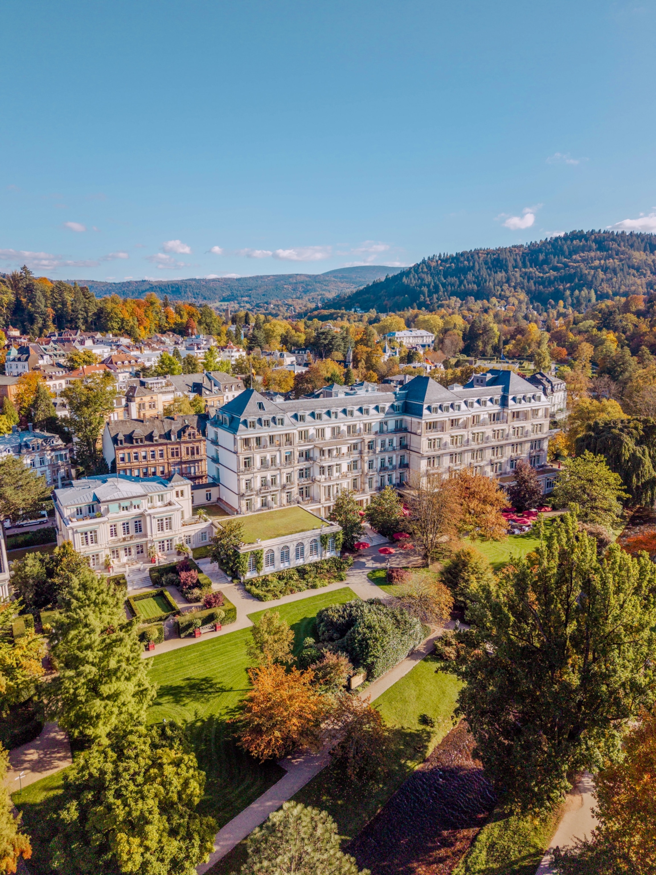 An aerial view of a grand hotel set among landscaped gardens, with a town and forested hills stretching into the distance.