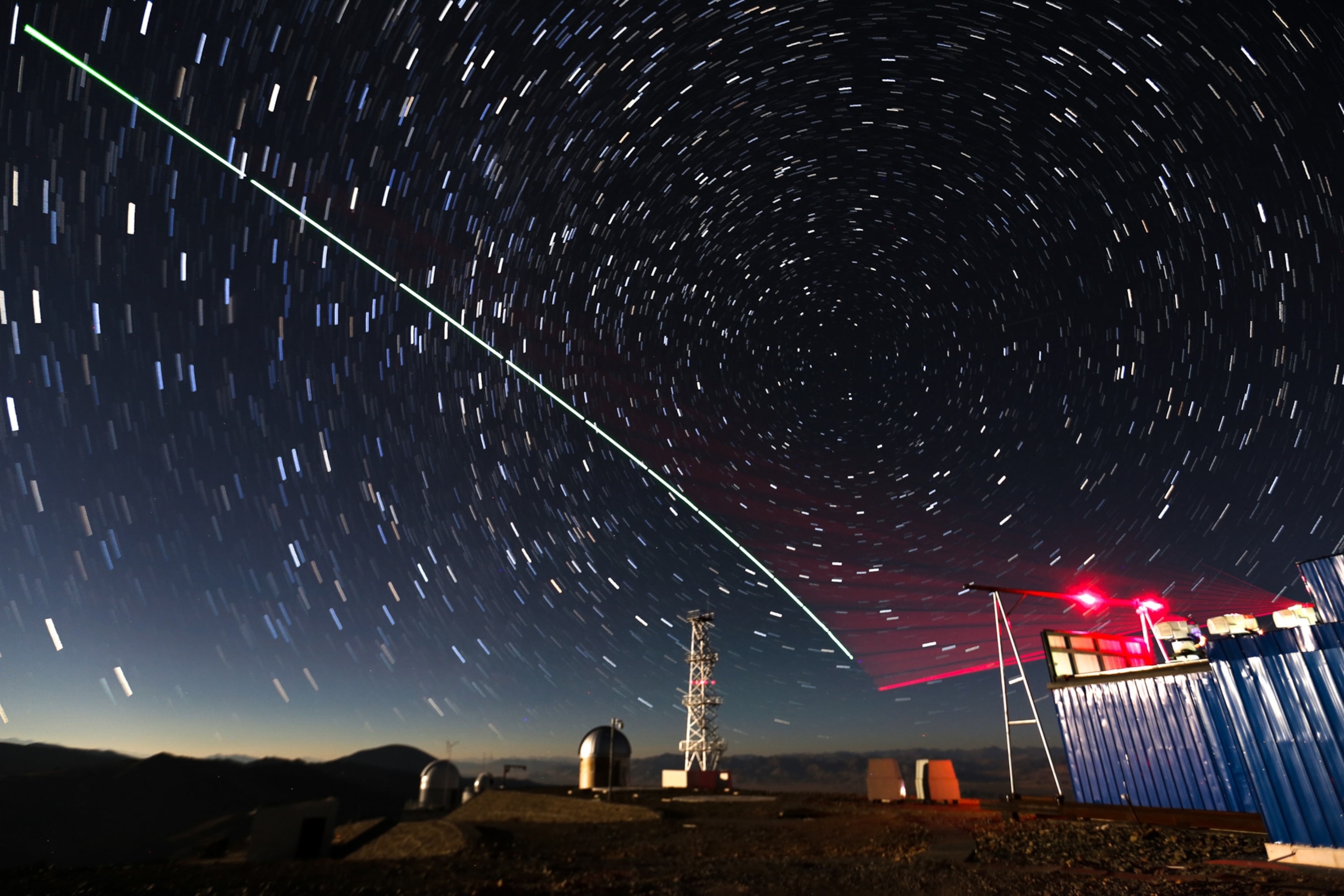 A starry night sky with swirling star trails and a bright green laser beam crossing diagonally. Below, observatory domes and a blue shed are visible.