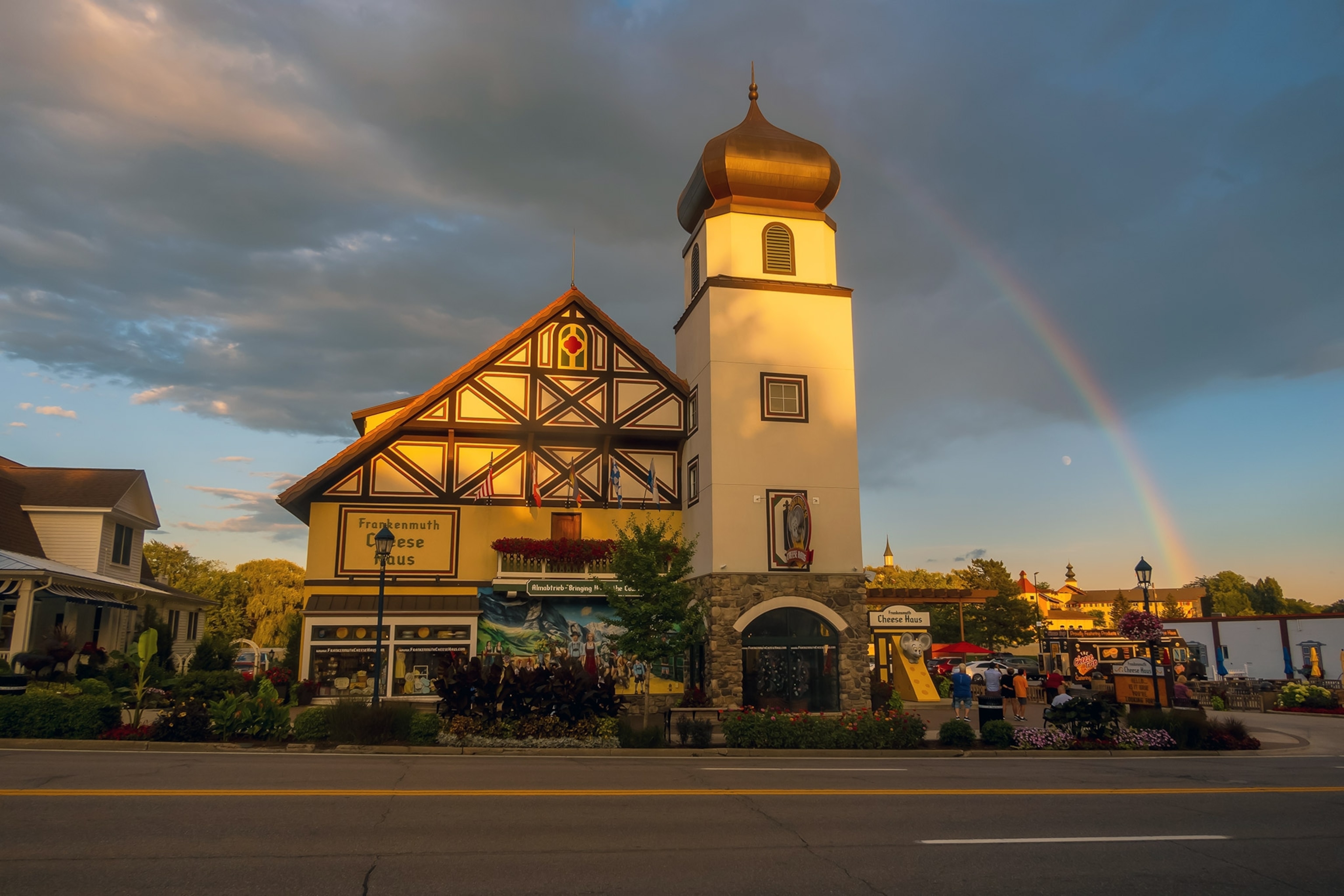 A European-style building with pitched roof and tower, sunlit, with dark sky above and a rainbow arcing over
