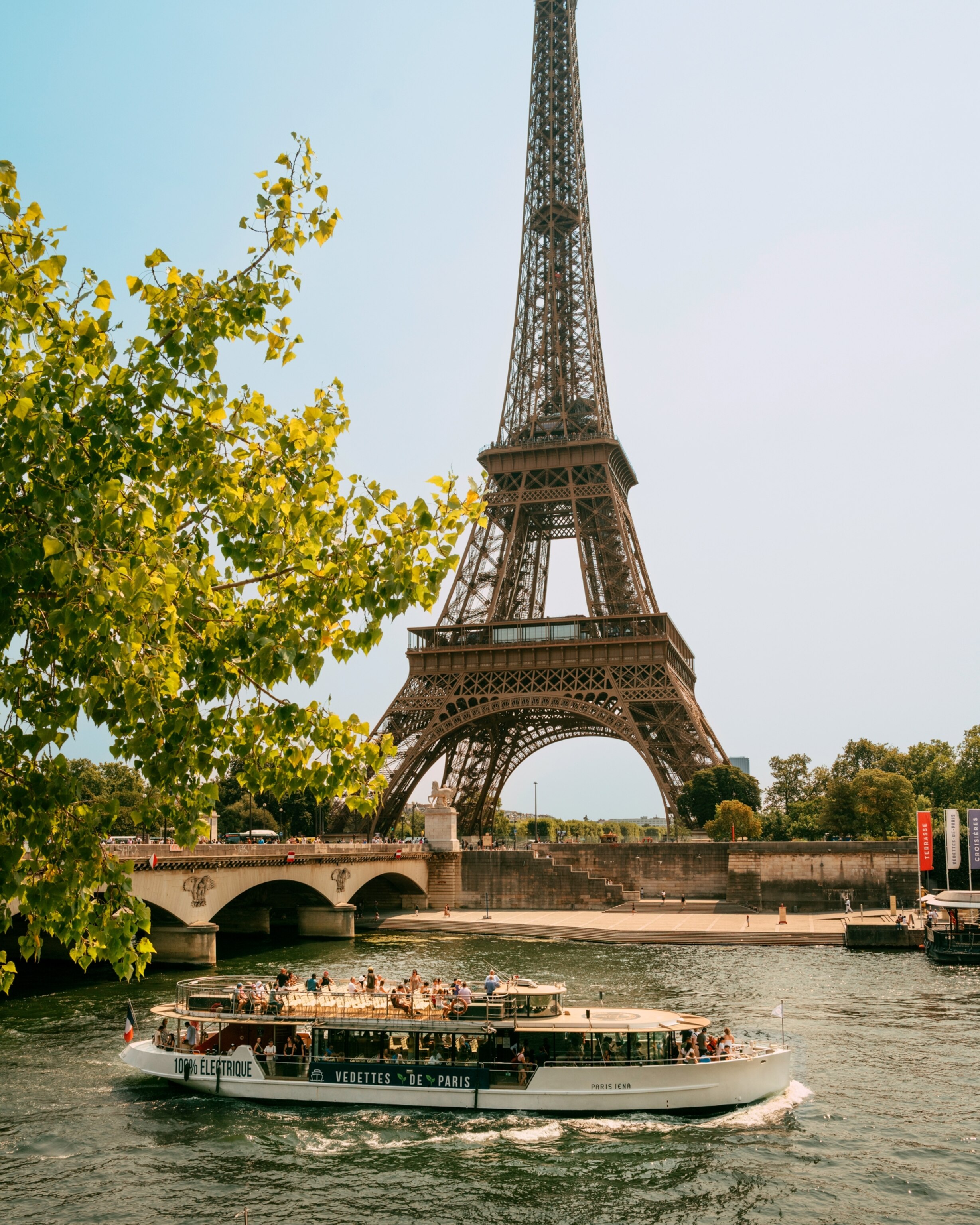The Eiffel Tower rises above the Seine River as a sightseeing boat passes below, with leafy trees framing the scene on a sunny day.