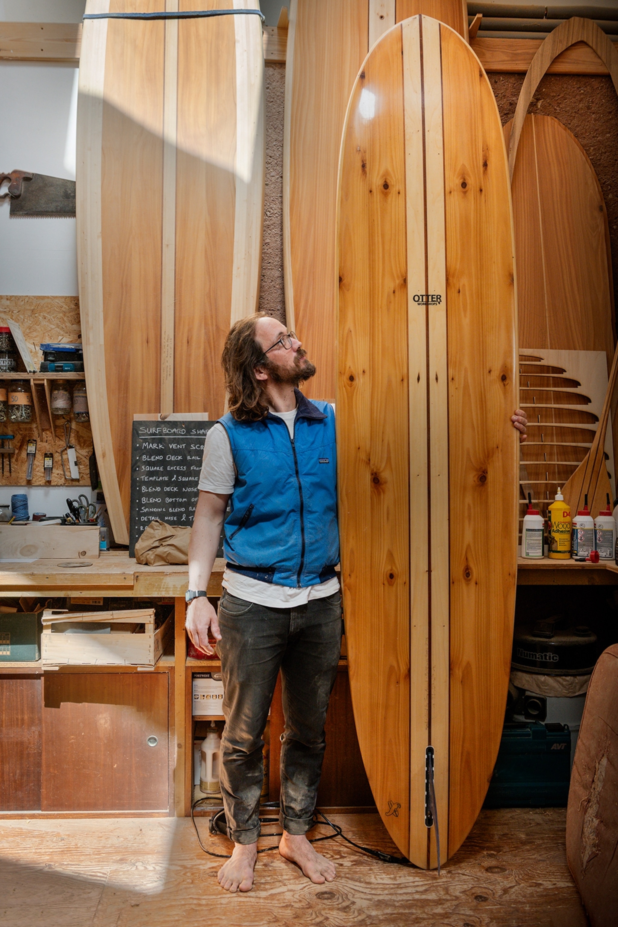 A young man standing next to a wooden surfboard inside his studio.
