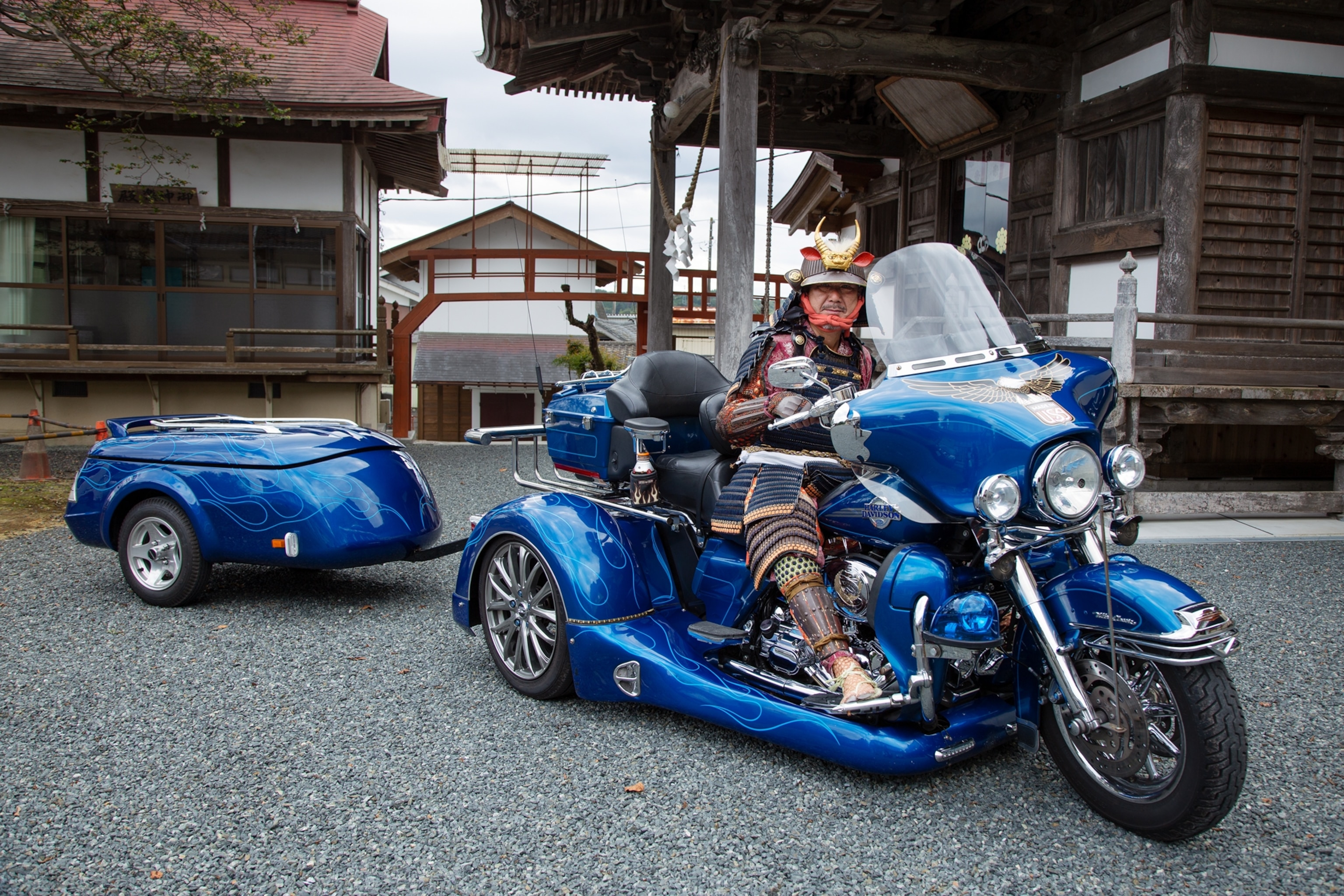 Picture of a man in samurai outfit on blue motorcycle with a trailer.
