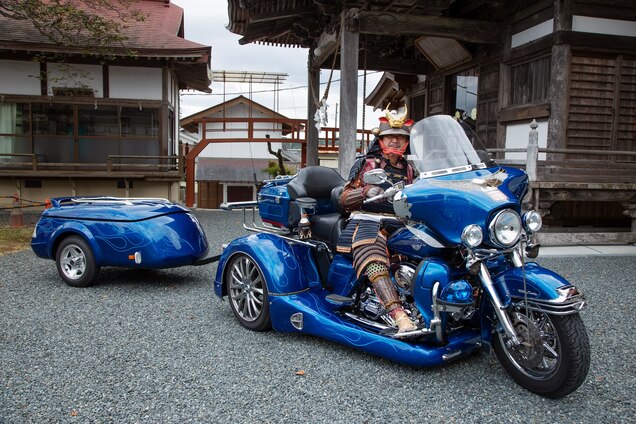 Picture of a man in samurai outfit on blue motorcycle with a trailer.