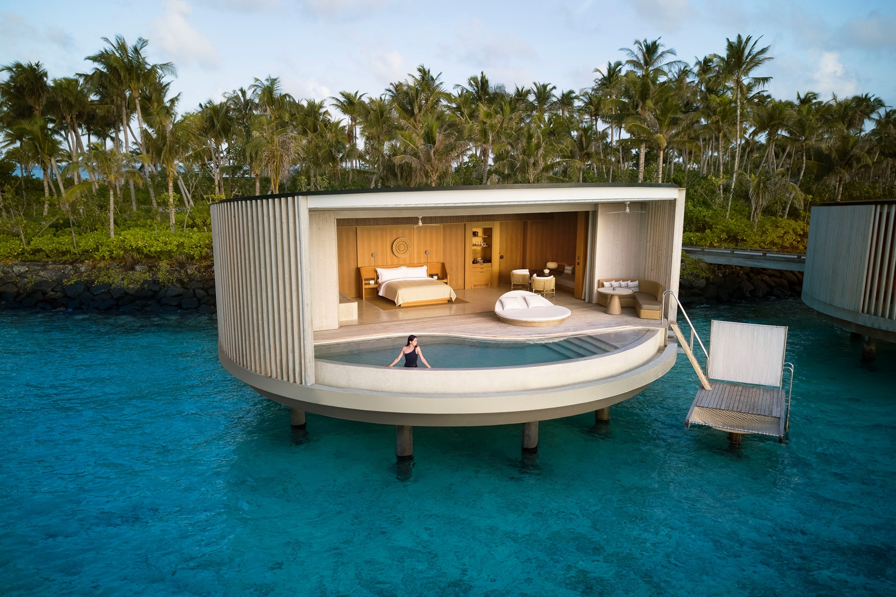 A drone shot of a circular, modern bungalow featuring an overwater pool and ocean-facing, open bedroom.