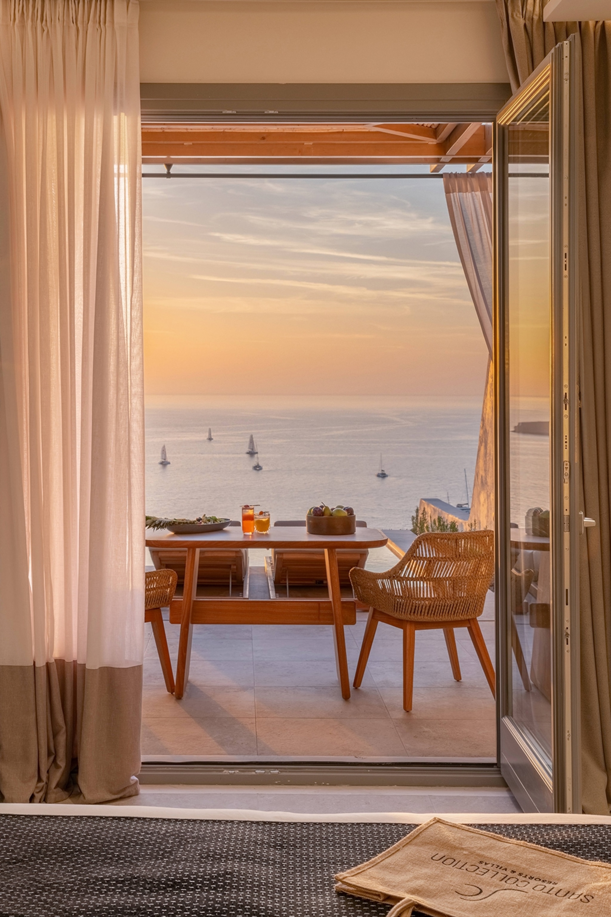 The interiors of a chic hotel bedroom with open double doors leading onto a terrace with a table and chairs at sunset.