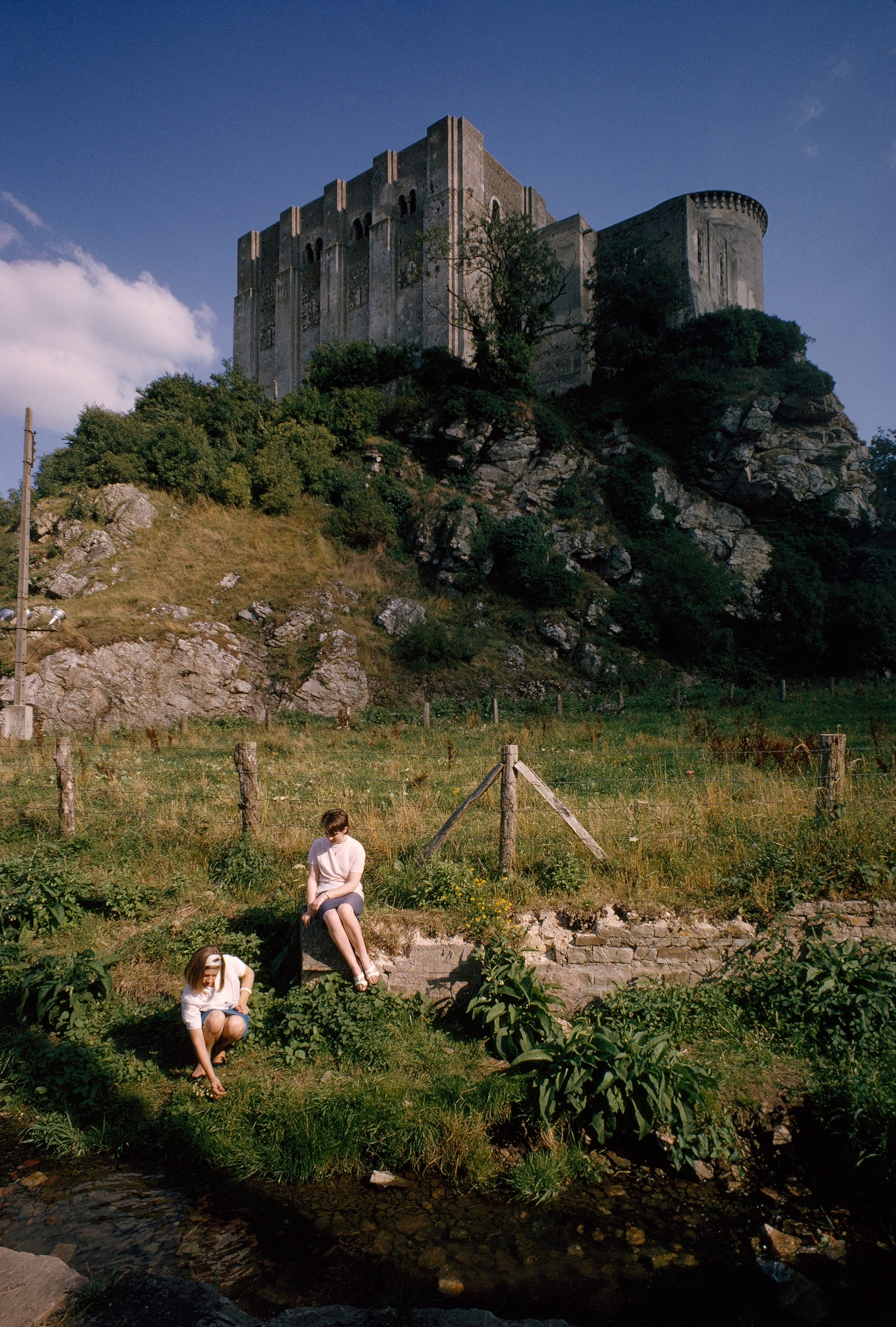 Falaise castle overlooks rocks, verdant pasture, and women at stream.