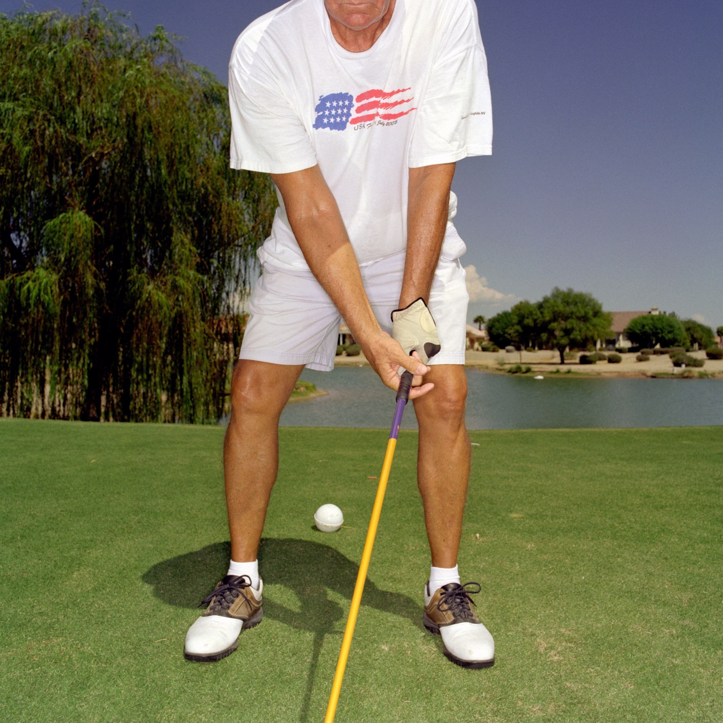 An elderly golfer in a white shirt and shorts stands on a green course, holding a yellow club with a golf ball nearby, with a pond and trees in the back.
