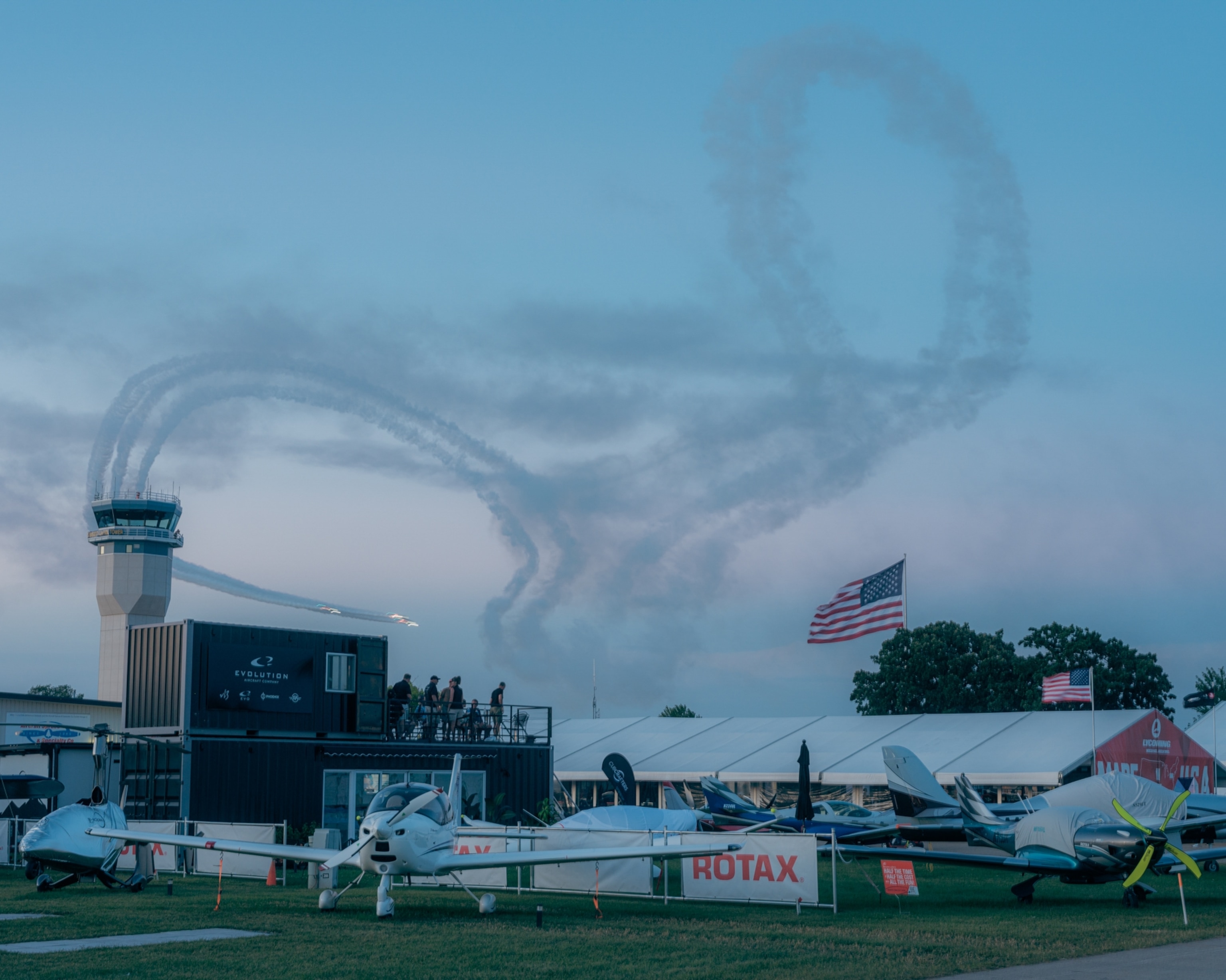 Airplanes perform a smoke trail display in a clear blue sky, forming loops and curves. Grounded aircraft, a control tower, and an American flag are visible below