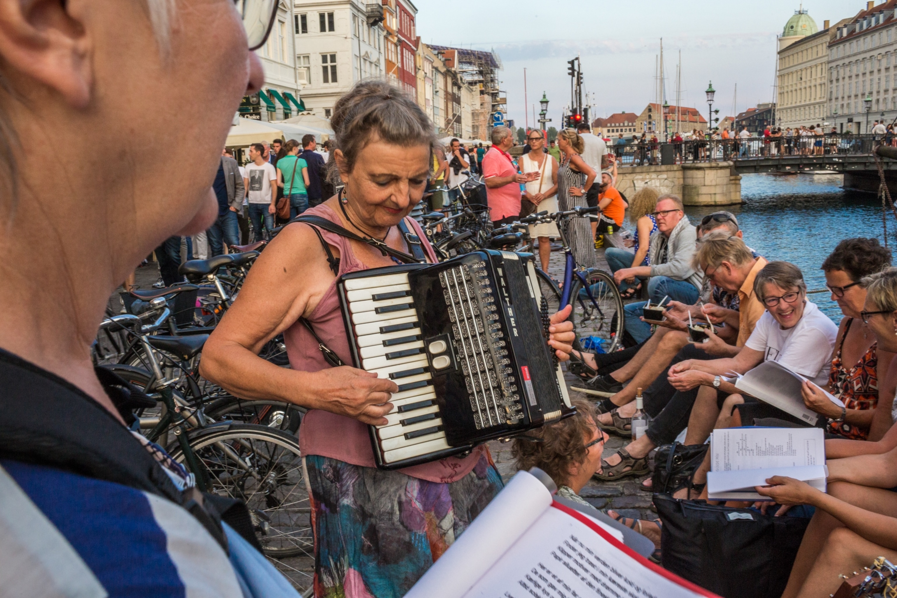 Crowds of people fill the streets by a canal in Copenhagen. In the foreground a group plays music, with a woman prominently seen as she plays accordion.