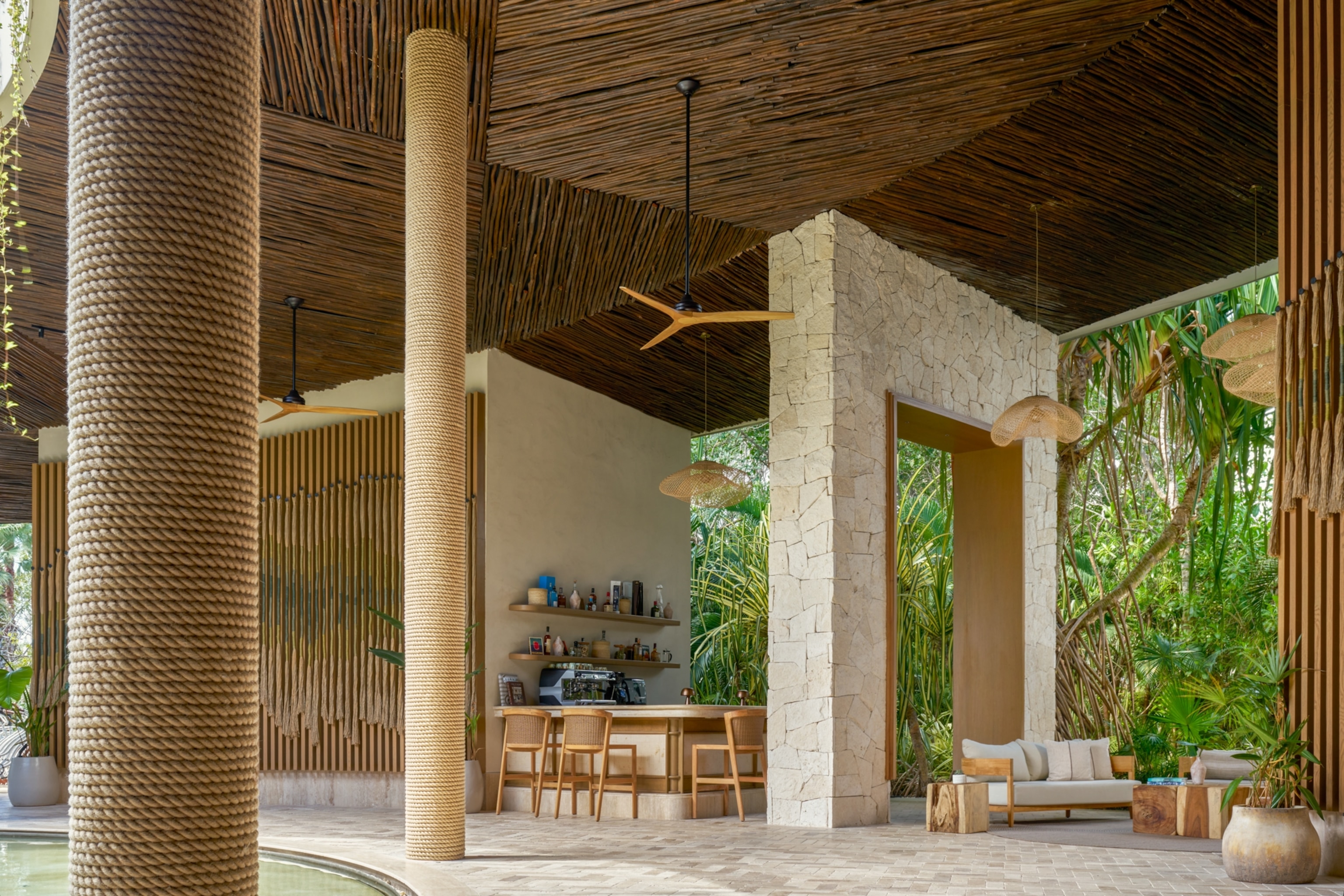 An open-air pavilion with rope-wrapped columns, natural wood and stone finishes, lounge seating, and lush tropical plants surrounding the space.
