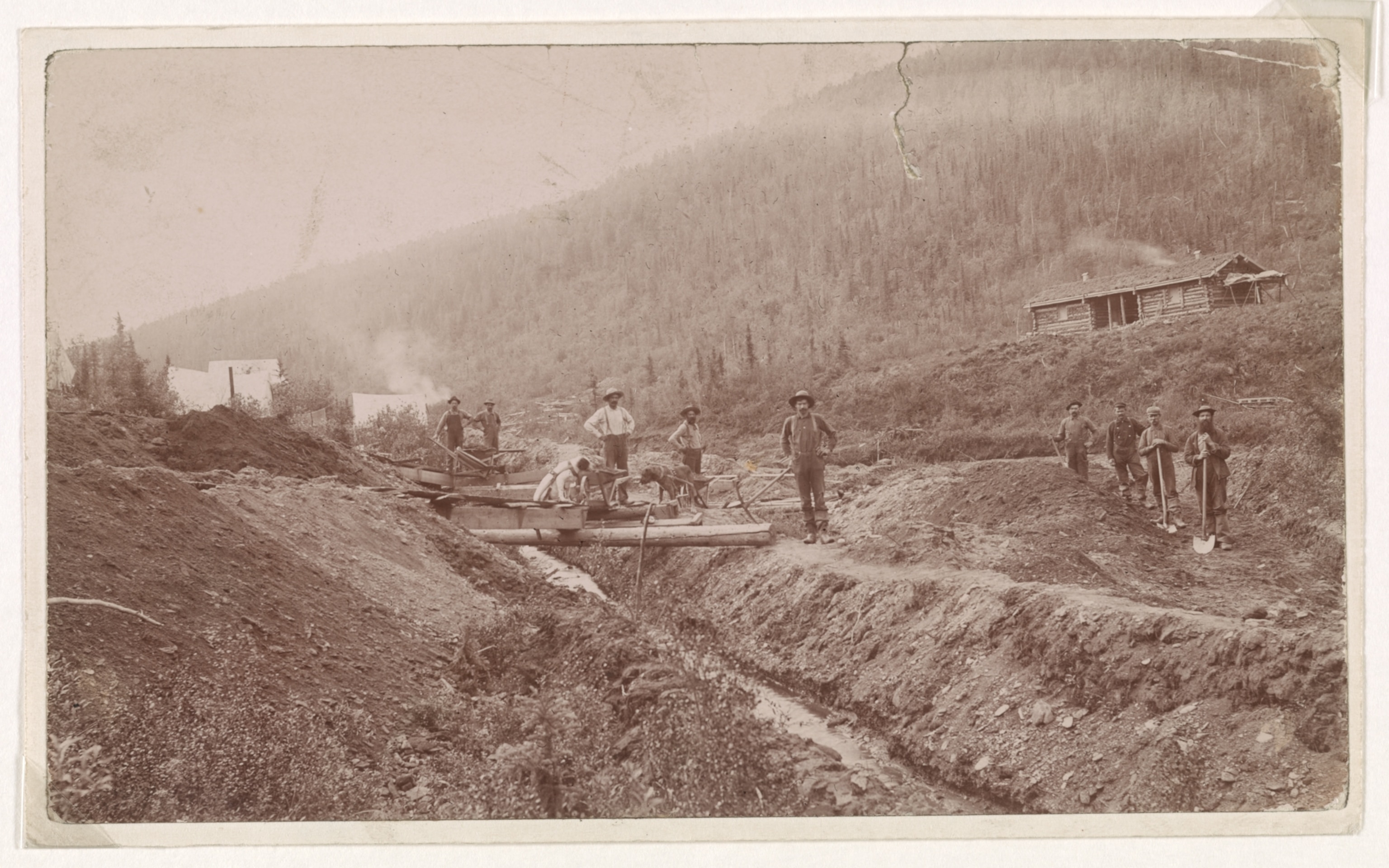 Photograph shows miner's camp and large log cabin on hillside; prospectors, some with shovels and pix axe pose near ditch and flume.