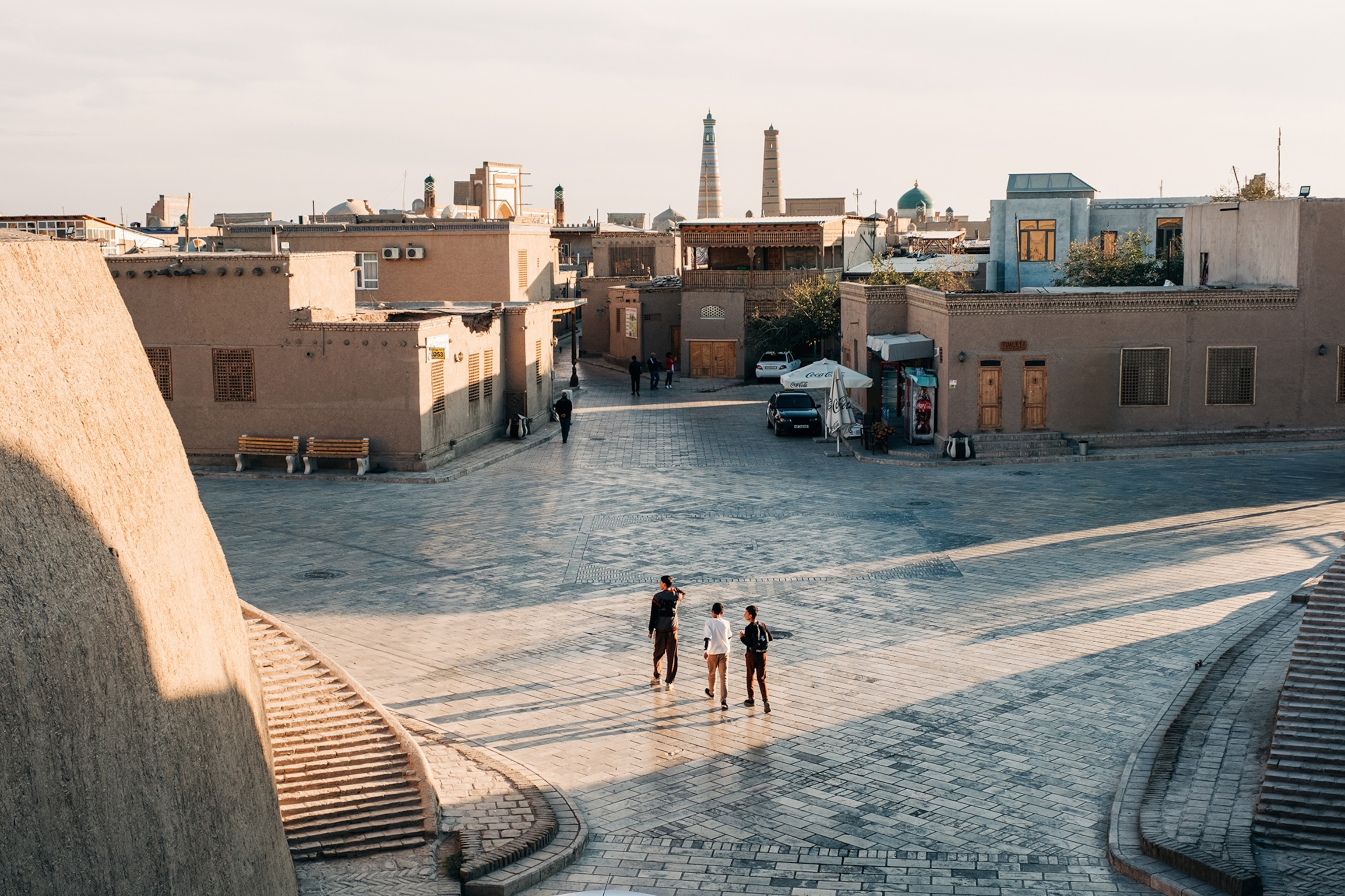 A late afternoon shot of an intersection in a low and flat-roofed medieval city with three visitors strolling around.