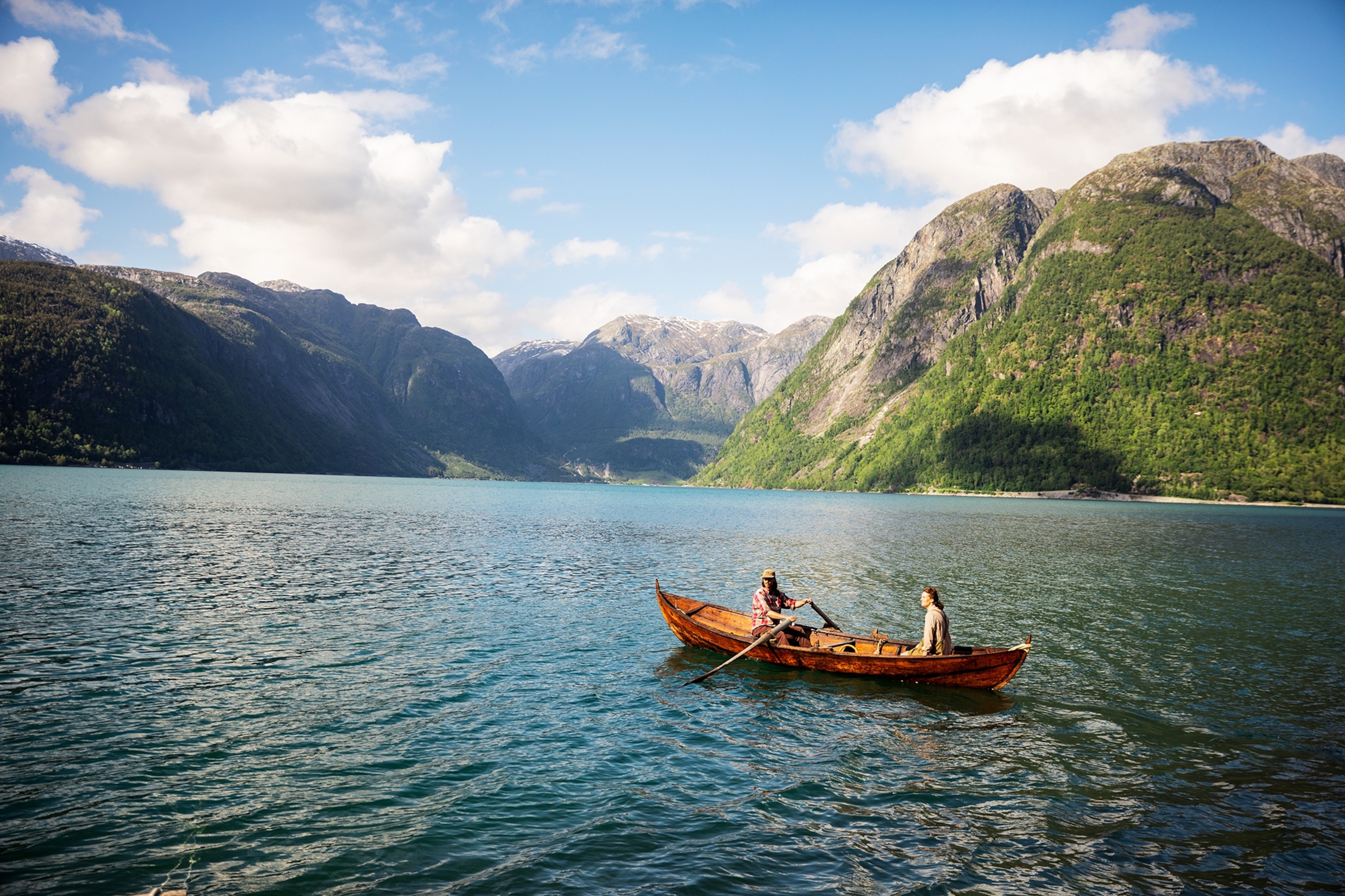 A small wooden rowboat carrying two people floats on a calm fjord, surrounded by steep green mountains and clear blue water under a partly cloudy sky.